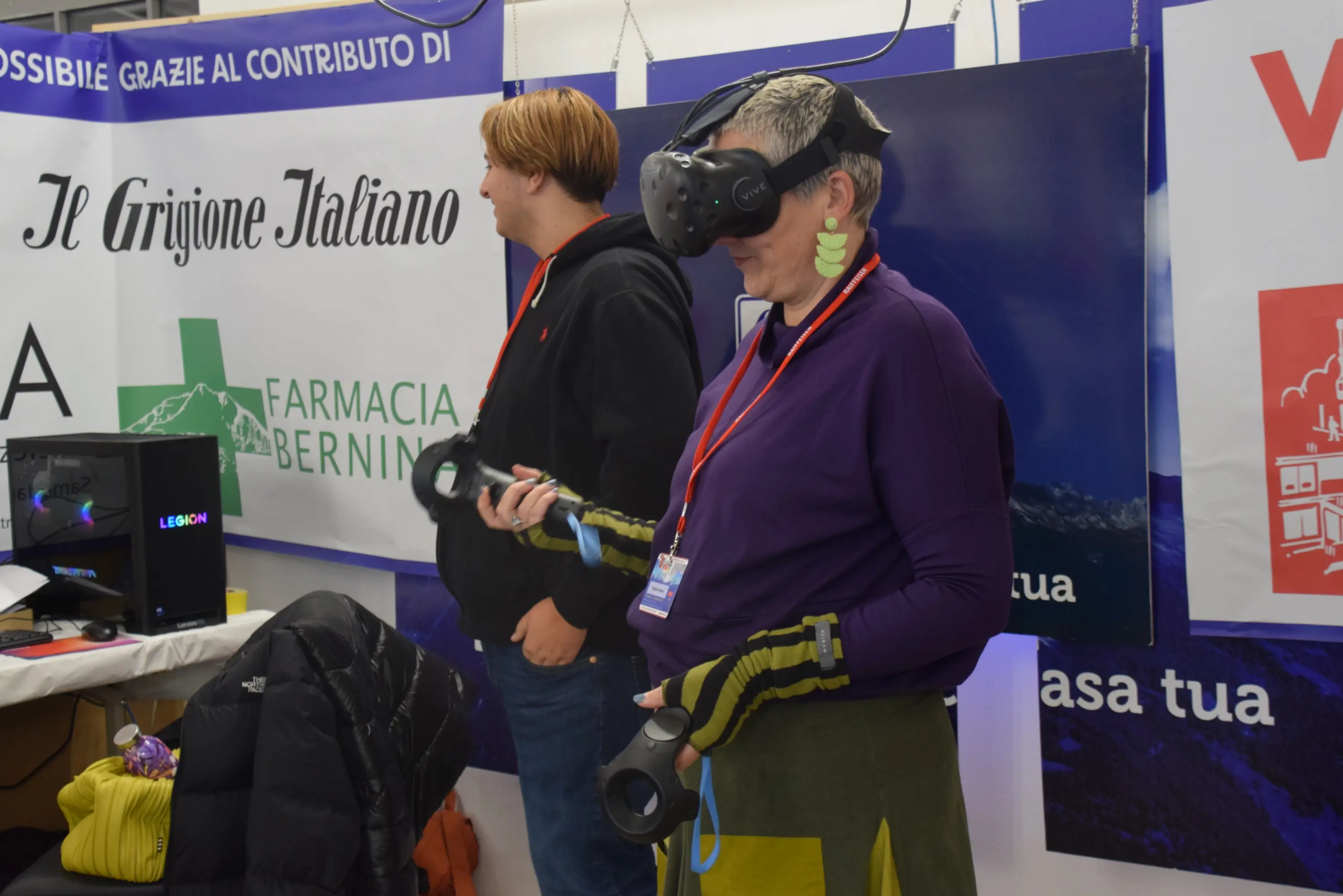 Due persone a un evento, una delle quali indossa un visore VR e tiene controller. Sullo sfondo, si vedono vari banner, tra cui uno con il logo di una farmacia. L'atmosfera è vivace e tecnologica.