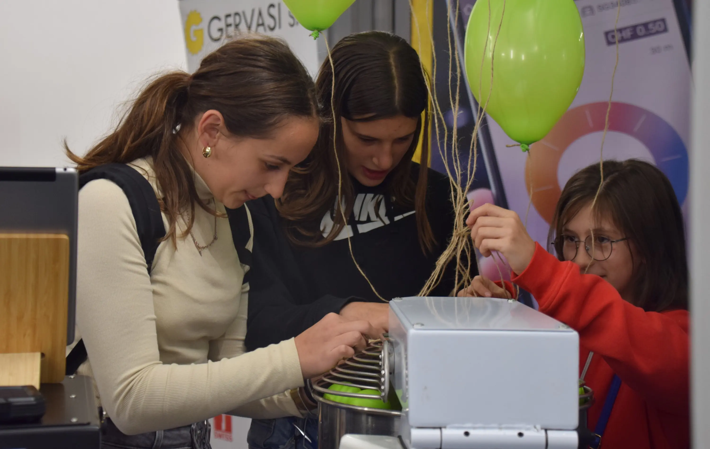 Tre ragazze interagiscono con un macchinario, mentre sono circondate da palloncini verdi. Si concentrano su quello che stanno facendo e mostrano un atteggiamento curioso e divertito.