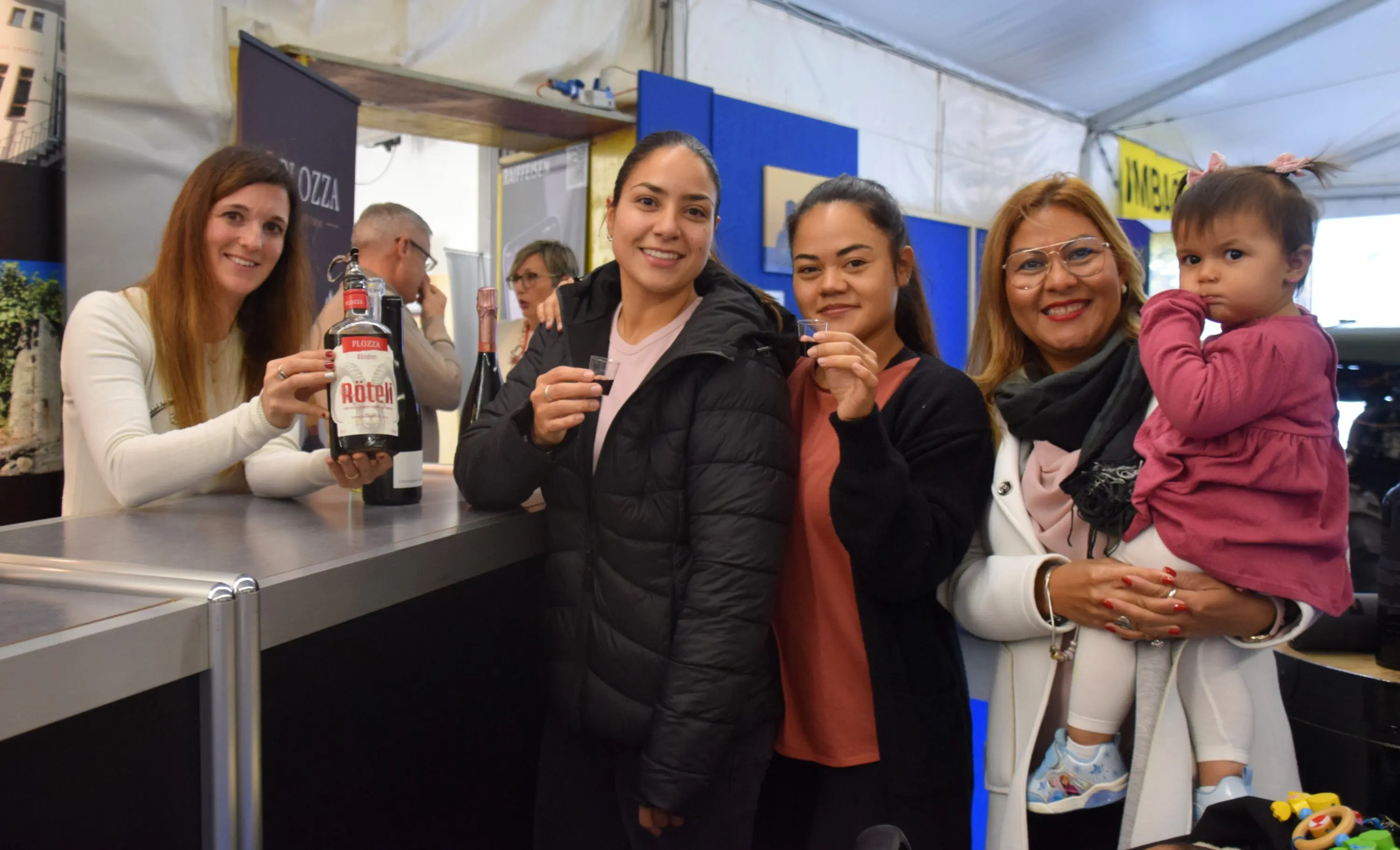 Quattro donne sorridenti in un evento, con una di loro che tiene in mano una bottiglia. Una donna tiene in braccio una bambina. Sullo sfondo, altre persone e stand espositivi.