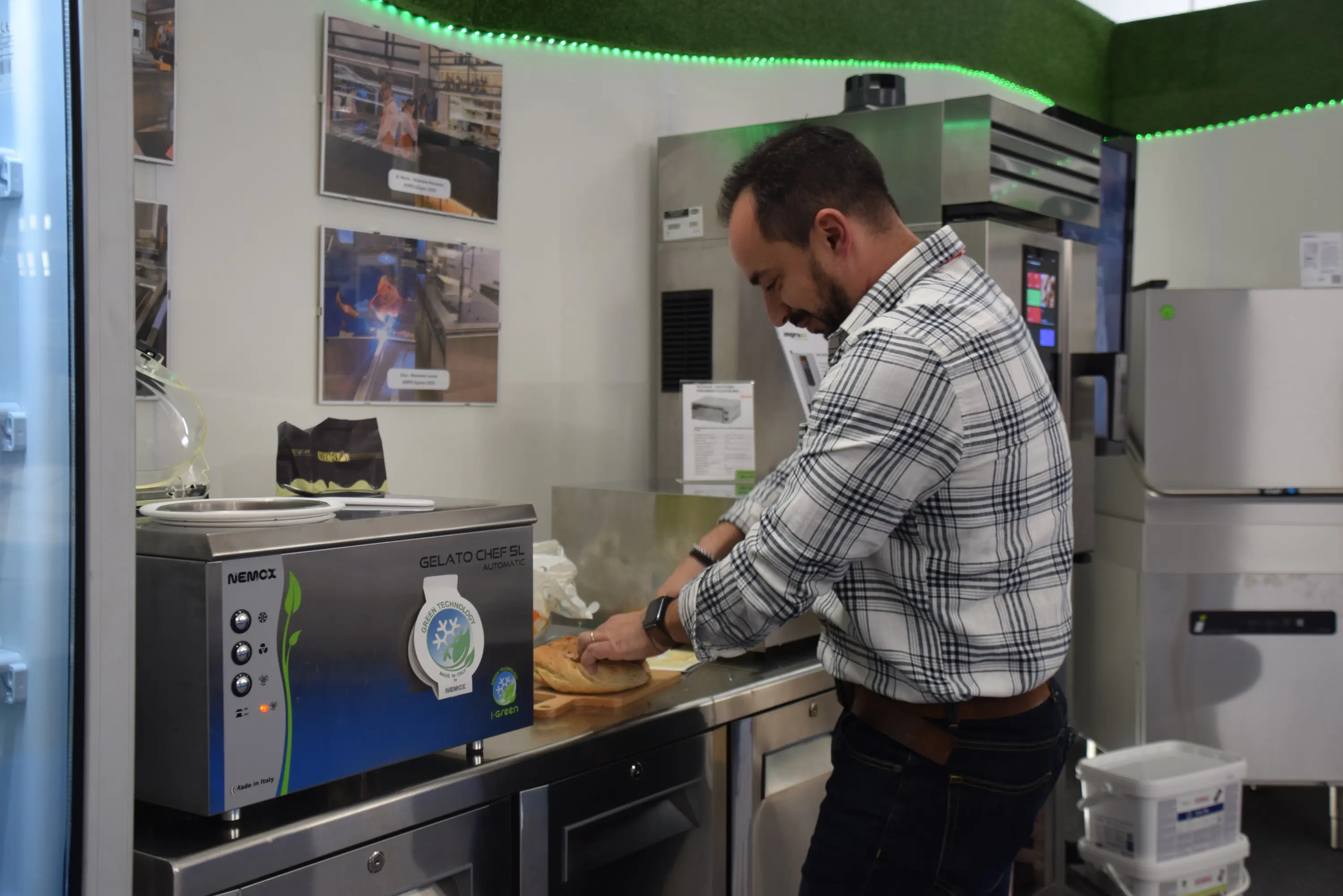 Un uomo sta preparando gelato in una cucina attrezzata, utilizzando una macchina per gelato. Sullo sfondo si vedono attrezzi da cucina e fotografie appese alle pareti.