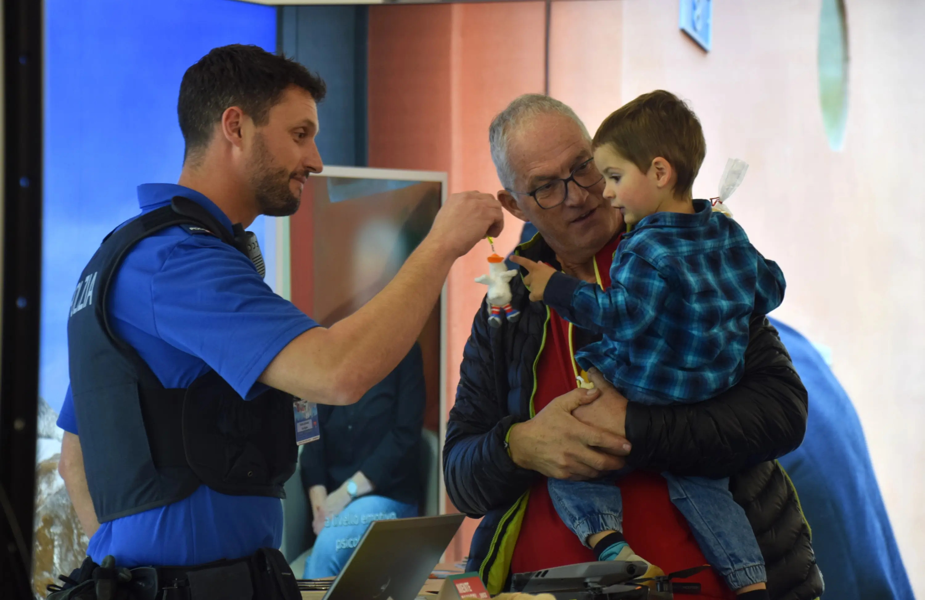 Un poliziotto interagisce con un bambino mentre un adulto, probabilmente il nonno, guarda con interesse. Il bambino tiene in mano un piccolo giocattolo.