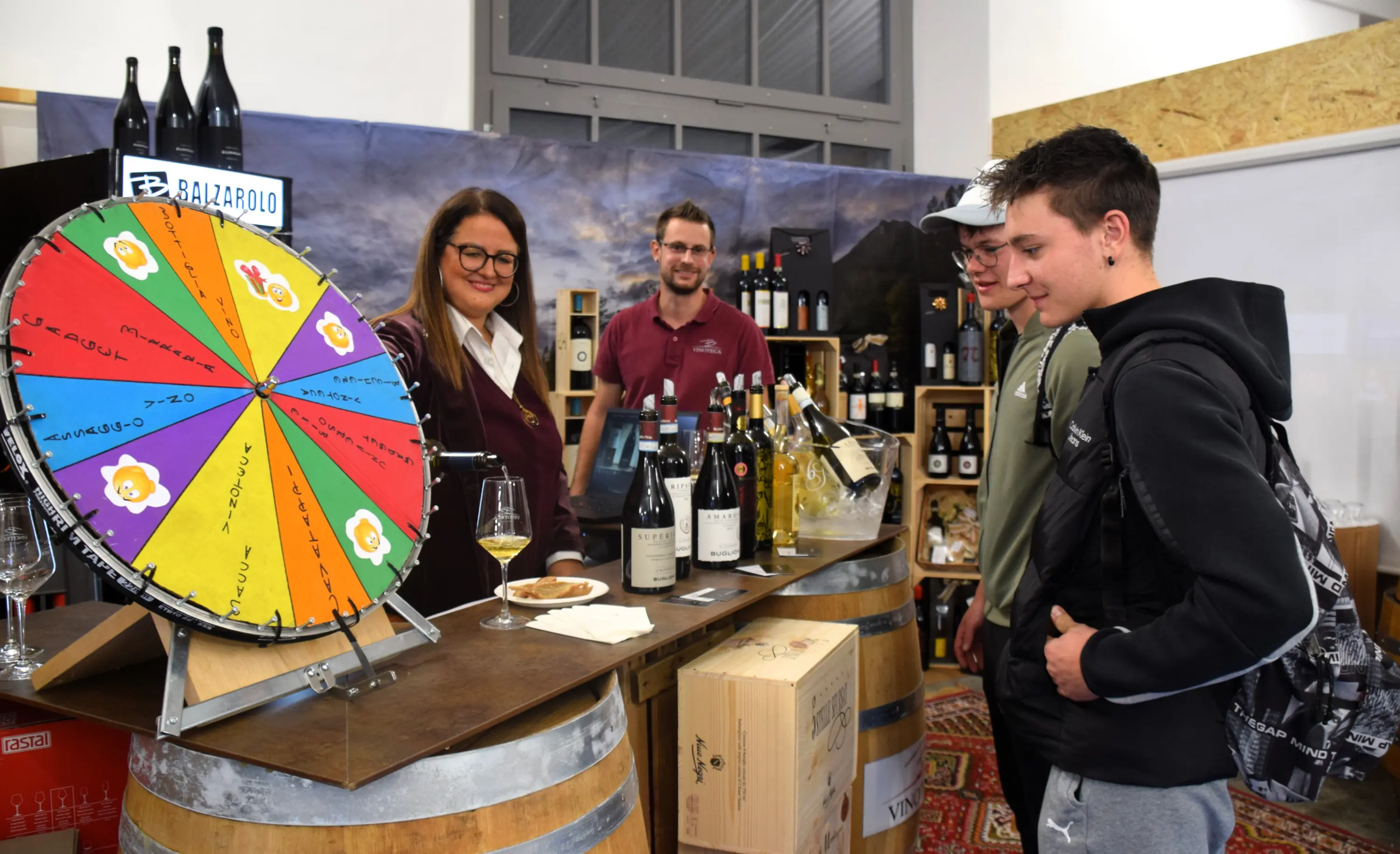 Una donna sorride mentre indica una ruota colorata a tre uomini in una fiera del vino. Sul tavolo ci sono diverse bottiglie di vino e un bicchiere sul lato. Ambiente accogliente con botti di legno e scaffali pieni di vini sullo sfondo.