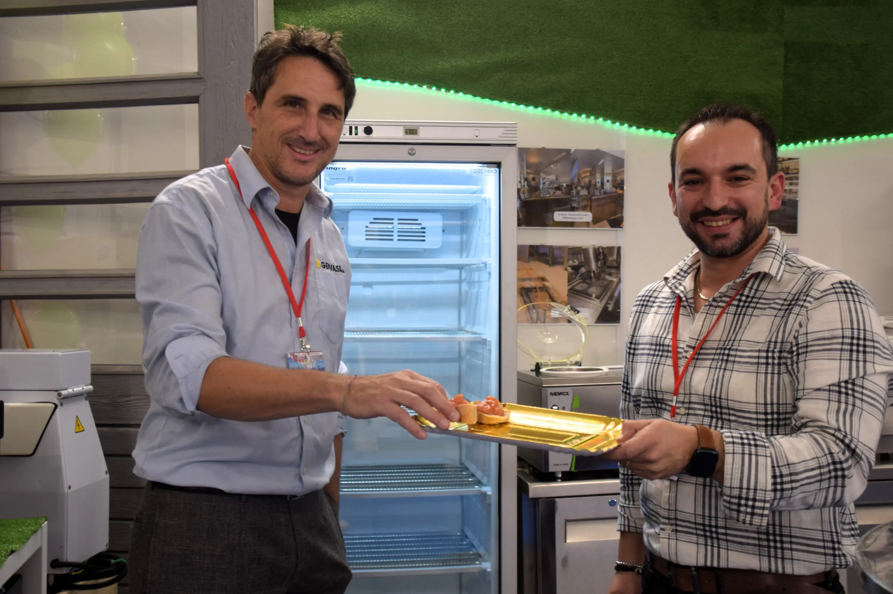 Due uomini sorridenti, uno dei quali tiene un vassoio con dolcetti, davanti a un frigorifero aperto. Entrambi indossano camicie e hanno distintivi al collo. Sullo sfondo, una parete con foto di progetti.