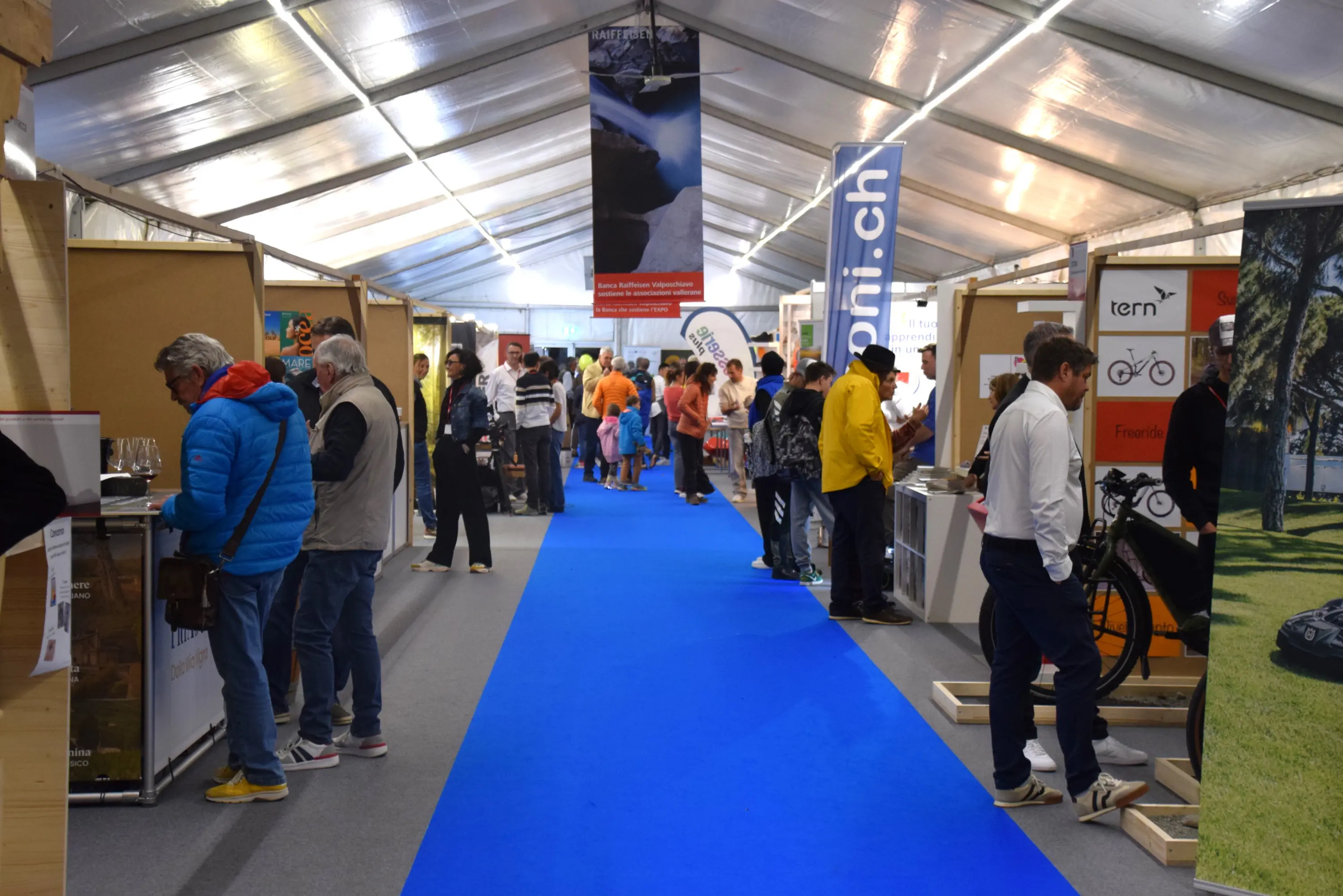 Una fiera con stand espositivi e una passerella blu. Persone stanno visitando gli stand, mentre alcune si fermano a osservare i prodotti in esposizione. L'atmosfera è vivace e affollata.