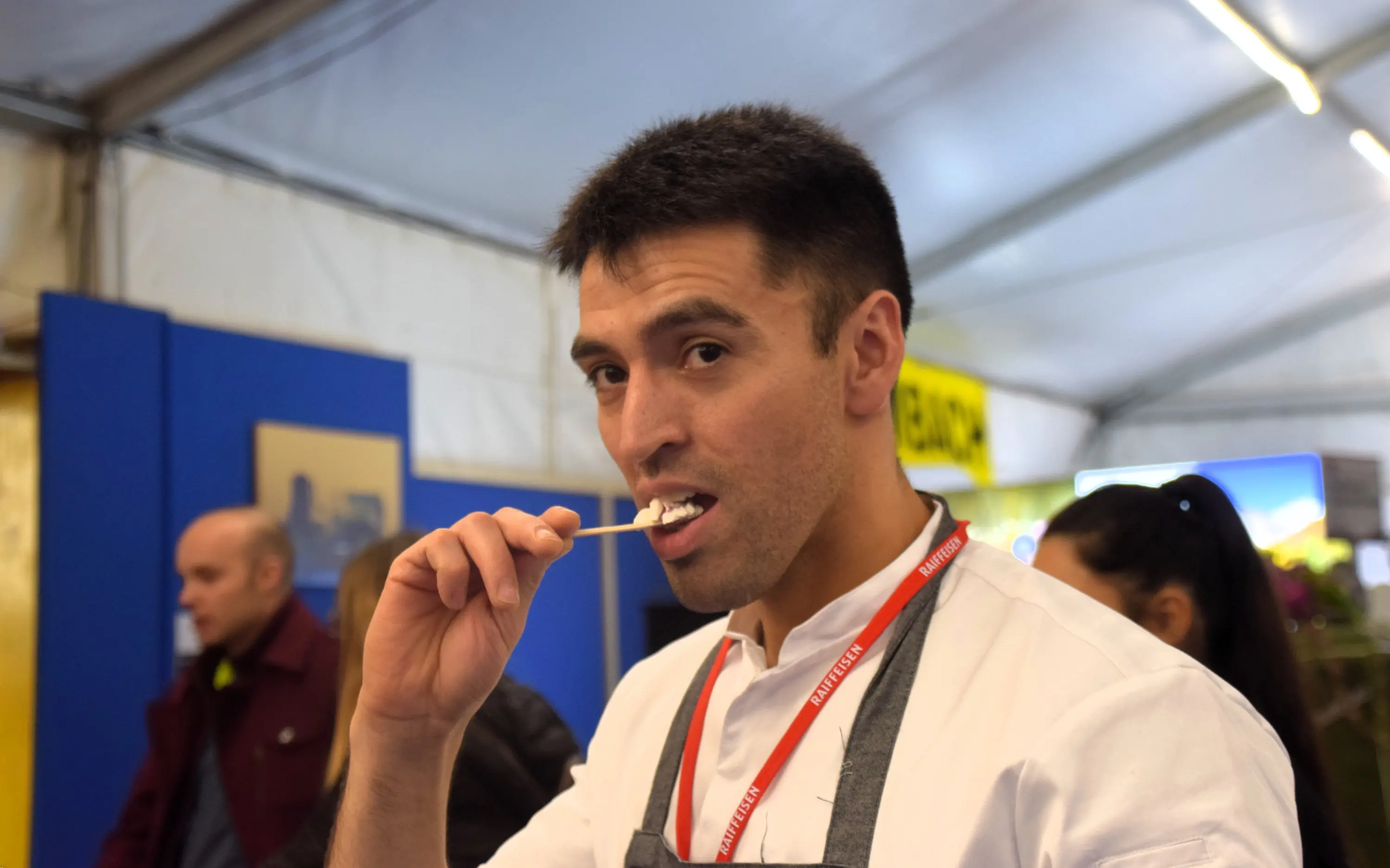 Un uomo con i capelli corti e un grembiule, che sorride mentre si spazzola i denti con uno spazzolino, all'interno di una tenda. Sullo sfondo si vedono altre persone e un ambiente di fiera.