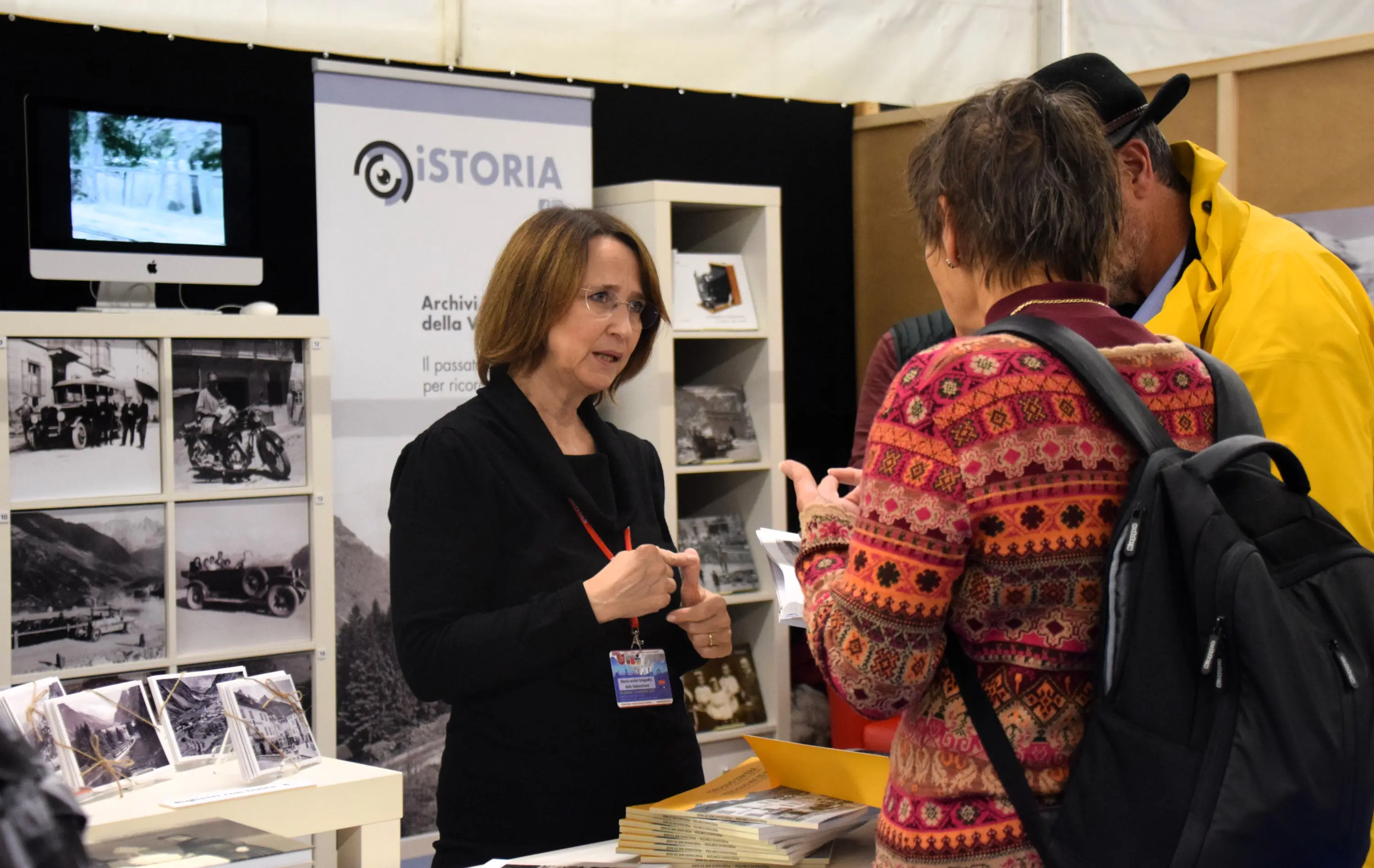 Una rappresentante di un archivio storico parla con due visitatori in uno stand espositivo. Alle spalle, ci sono fotografie storiche e un computer con uno schermo acceso.