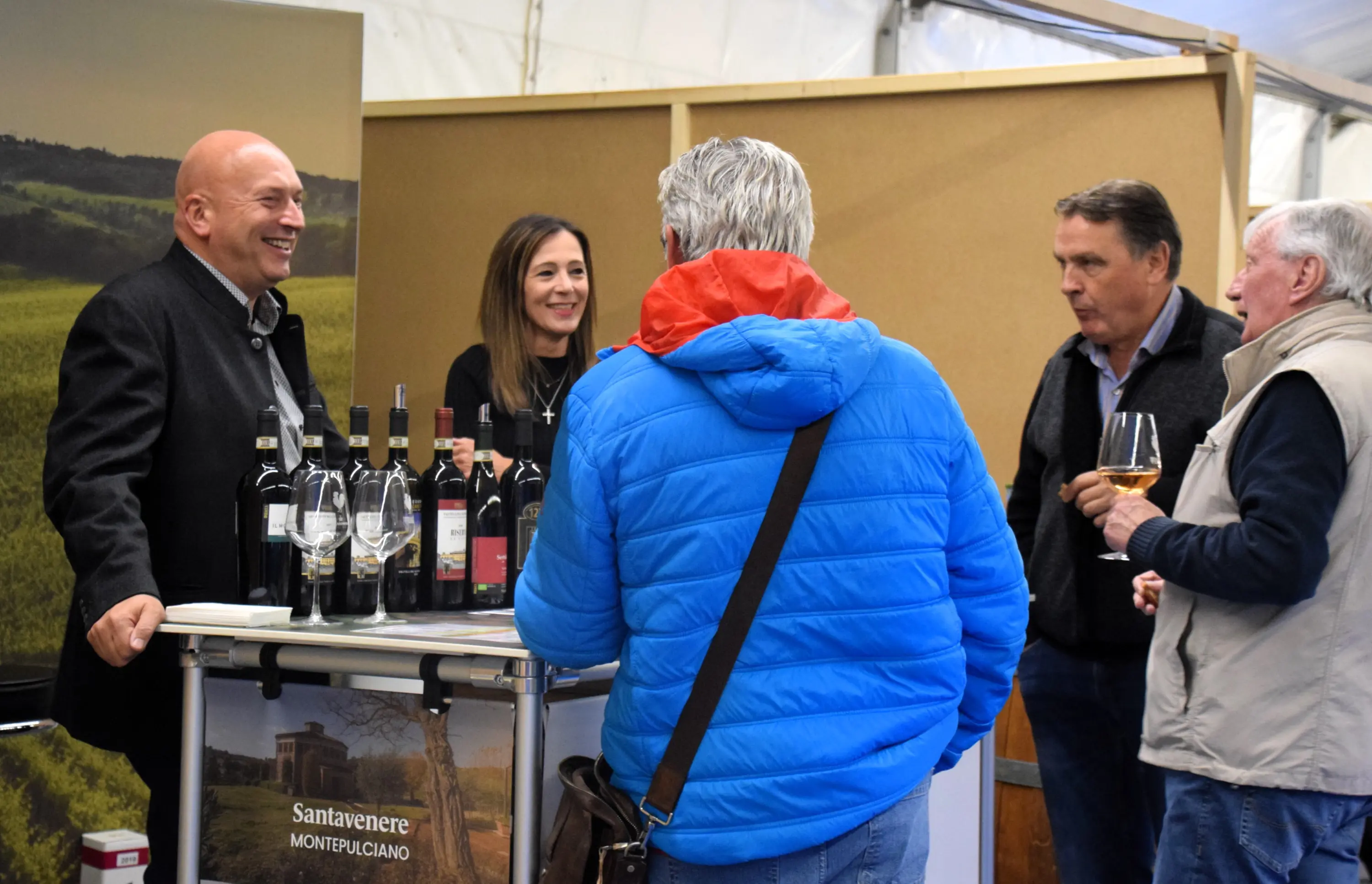 Un gruppo di quattro persone, di cui tre uomini e una donna, discutono davanti a un bancone di vino. Sono presenti bottiglie di vino esposte. Una delle persone tiene un bicchiere di vino mentre sorridono e conversano. Sullo sfondo si vedono colline verdi.