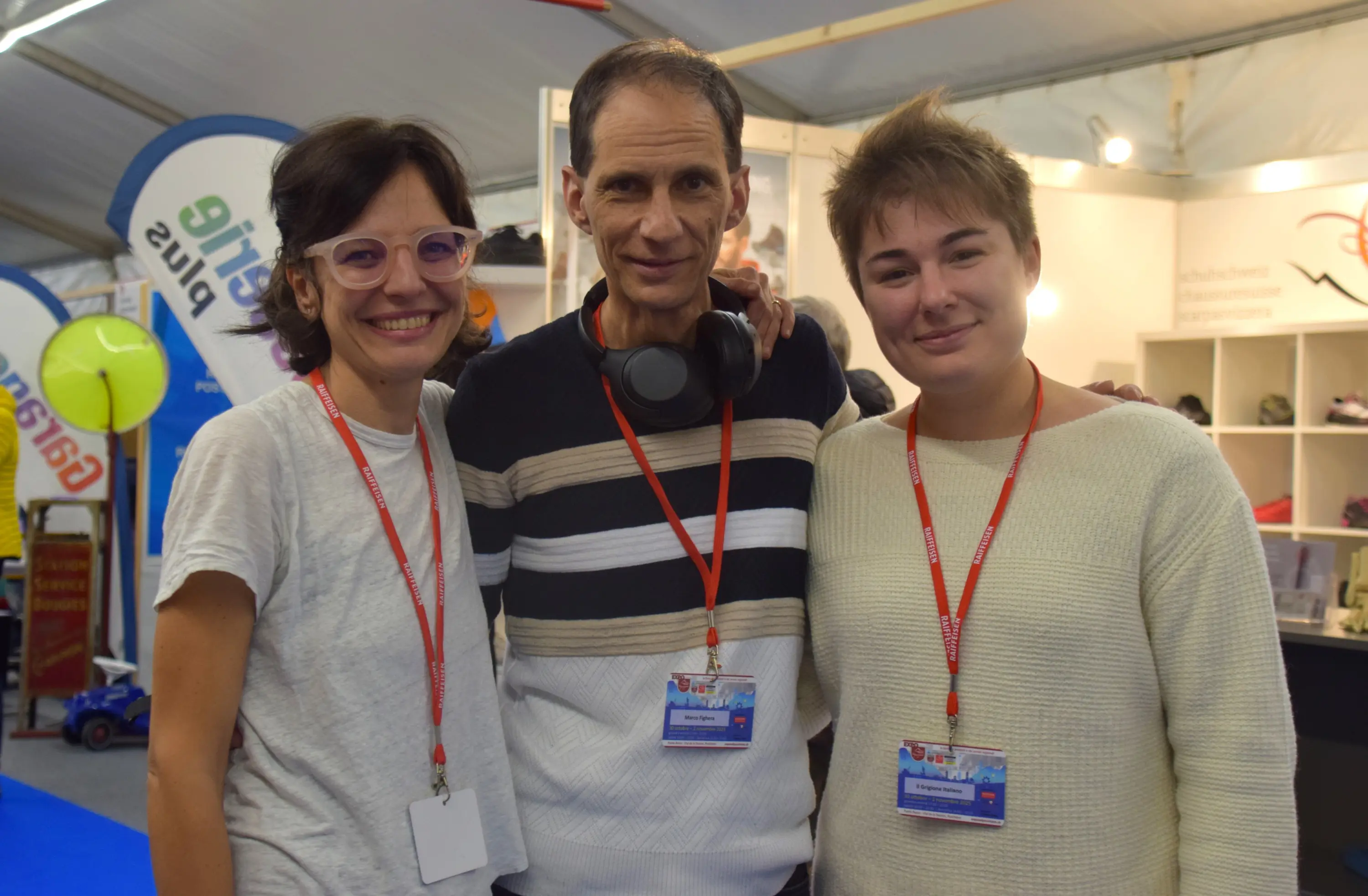Tre persone sorridenti si trovano in un ambiente interno, con poster e attrezzature sportive sullo sfondo. Due di loro indossano occhiali, mentre una ha le cuffie al collo. Tutti e tre indossano un pass identificativo.
