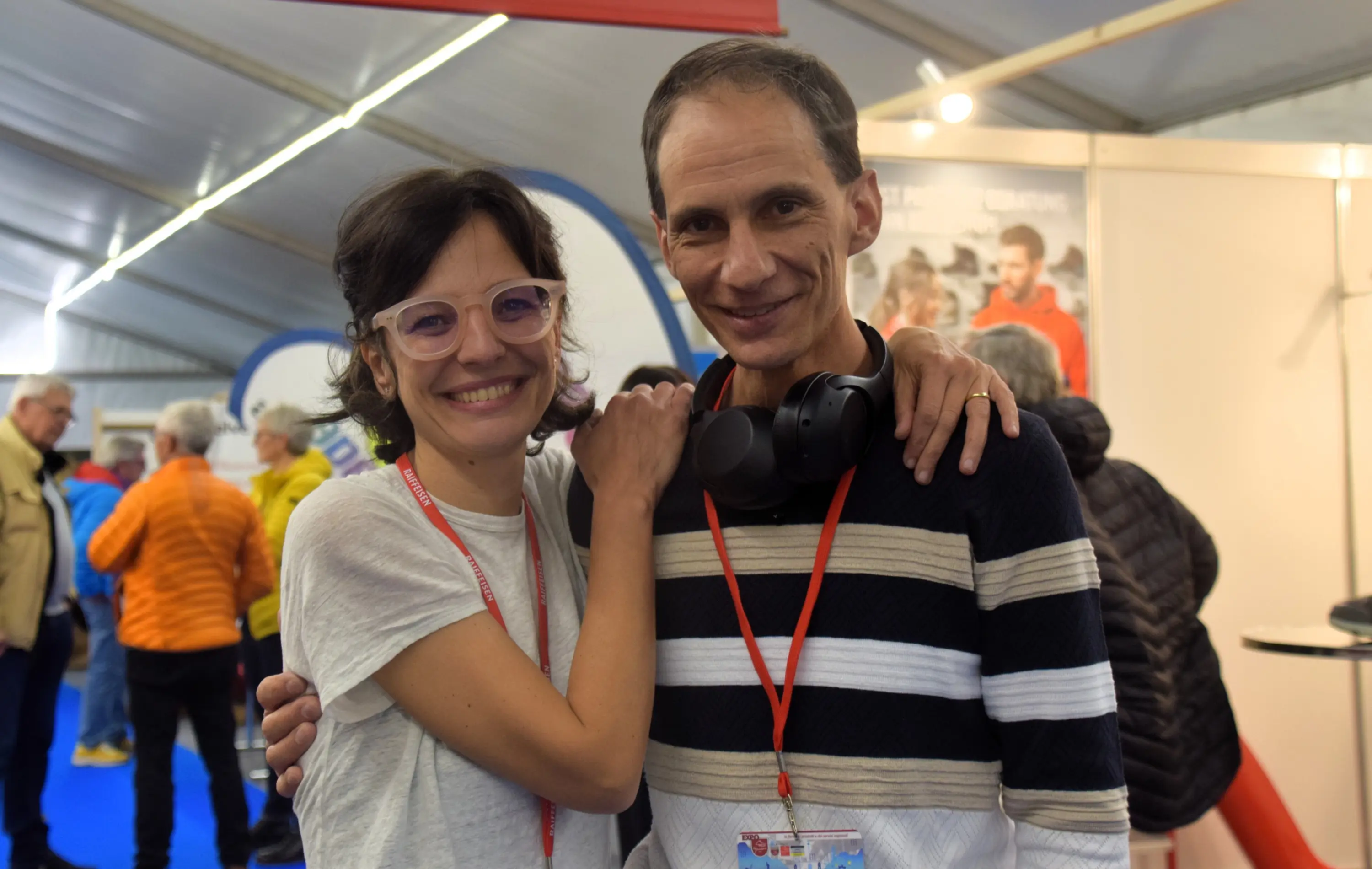 Due persone sorridenti, un uomo e una donna, si abbracciano mentre sono in un contesto di fiera con stand e visitatori sullo sfondo. La donna indossa occhiali chiari e una maglietta nera, mentre l'uomo ha cuffie intorno al collo e indossa una maglia a righe.