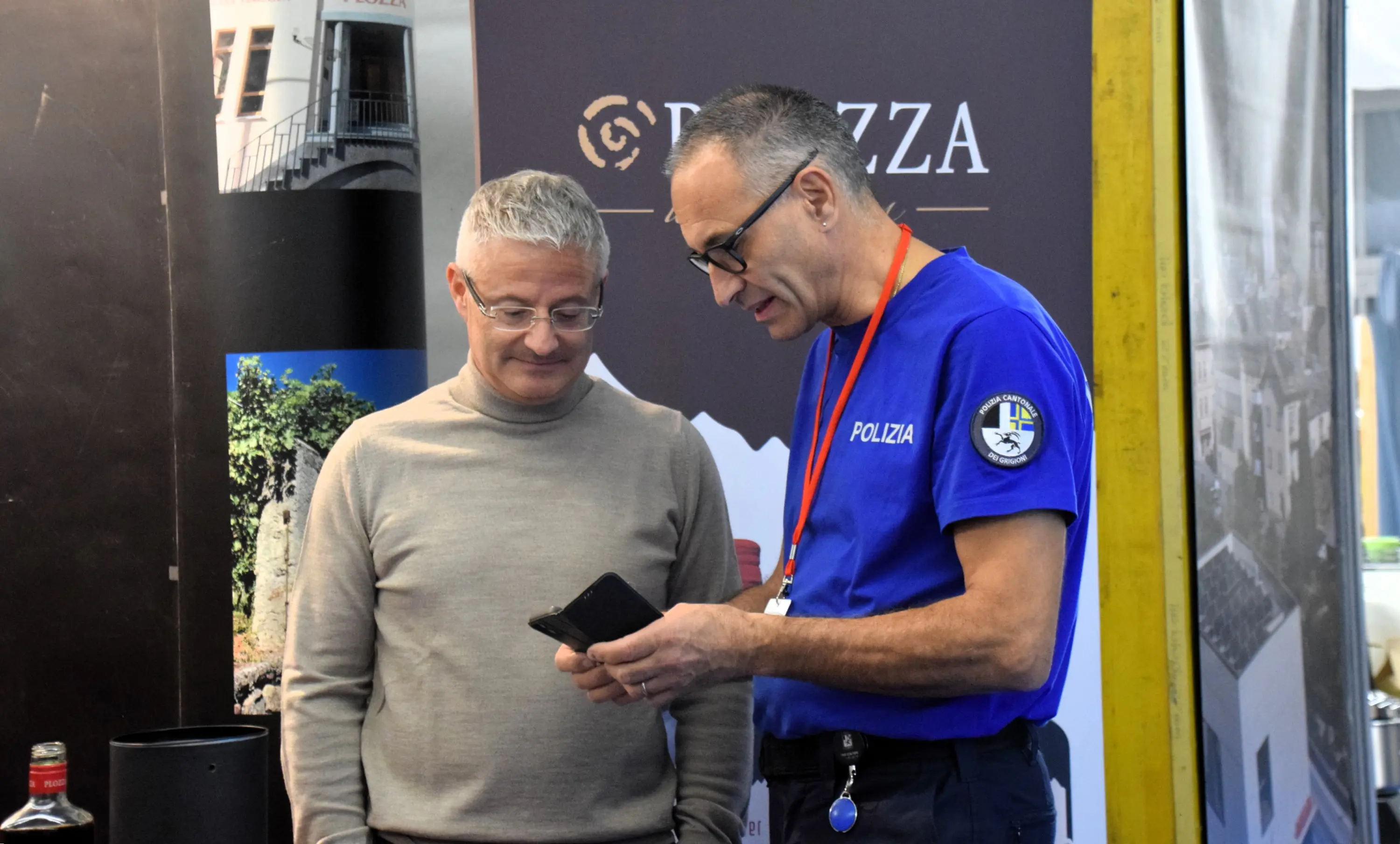 Due uomini stanno conversando a un evento, uno indossa un maglione beige e l'altro una maglietta blu con un distintivo. Entrambi guardano uno smartphone. Sullo sfondo, si vedono banner pubblicitari.
