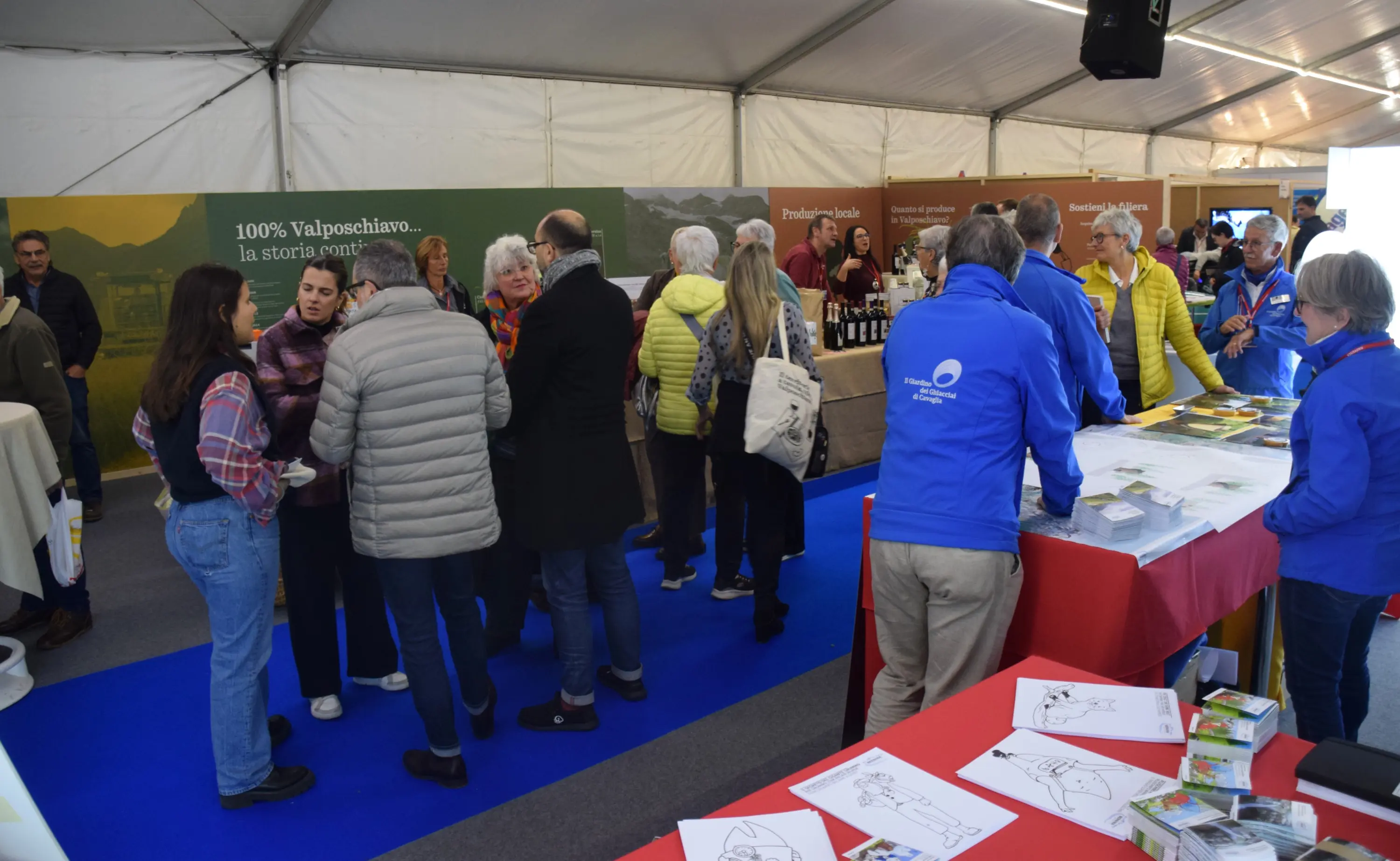 Un gruppo di persone si riunisce all'interno di una tenda a un evento, con stand espositivi che presentano prodotti locali. Alcuni partecipanti conversano animatamente, mentre altri esplorano i materiali su un tavolo. L'atmosfera è vivace e coinvolgente.