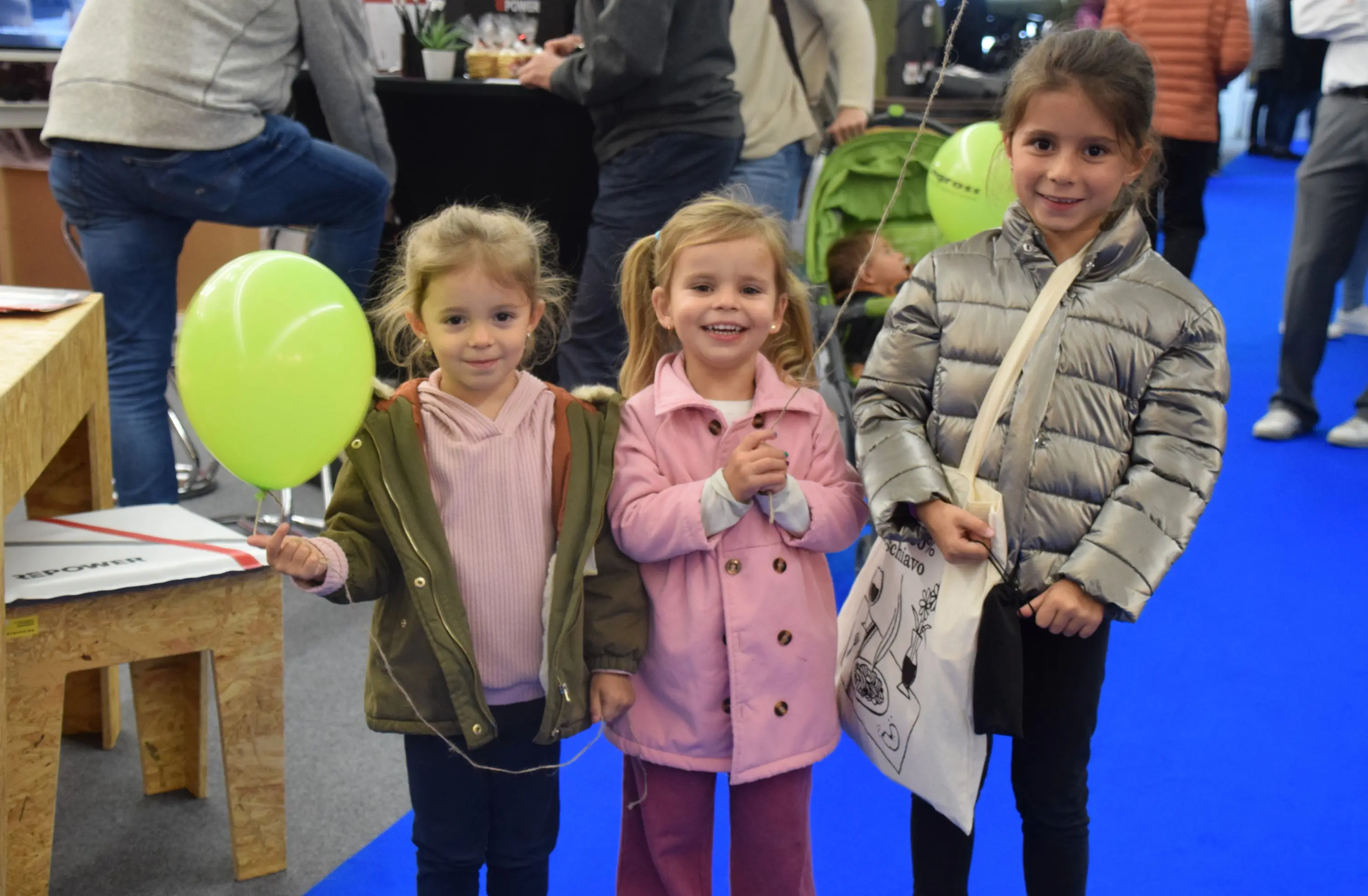 Tre bambine sorridenti in un ambiente festoso, con una delle ragazze che tiene un palloncino verde. Indossano giubbini colorati e si trovano su un tappeto blu.