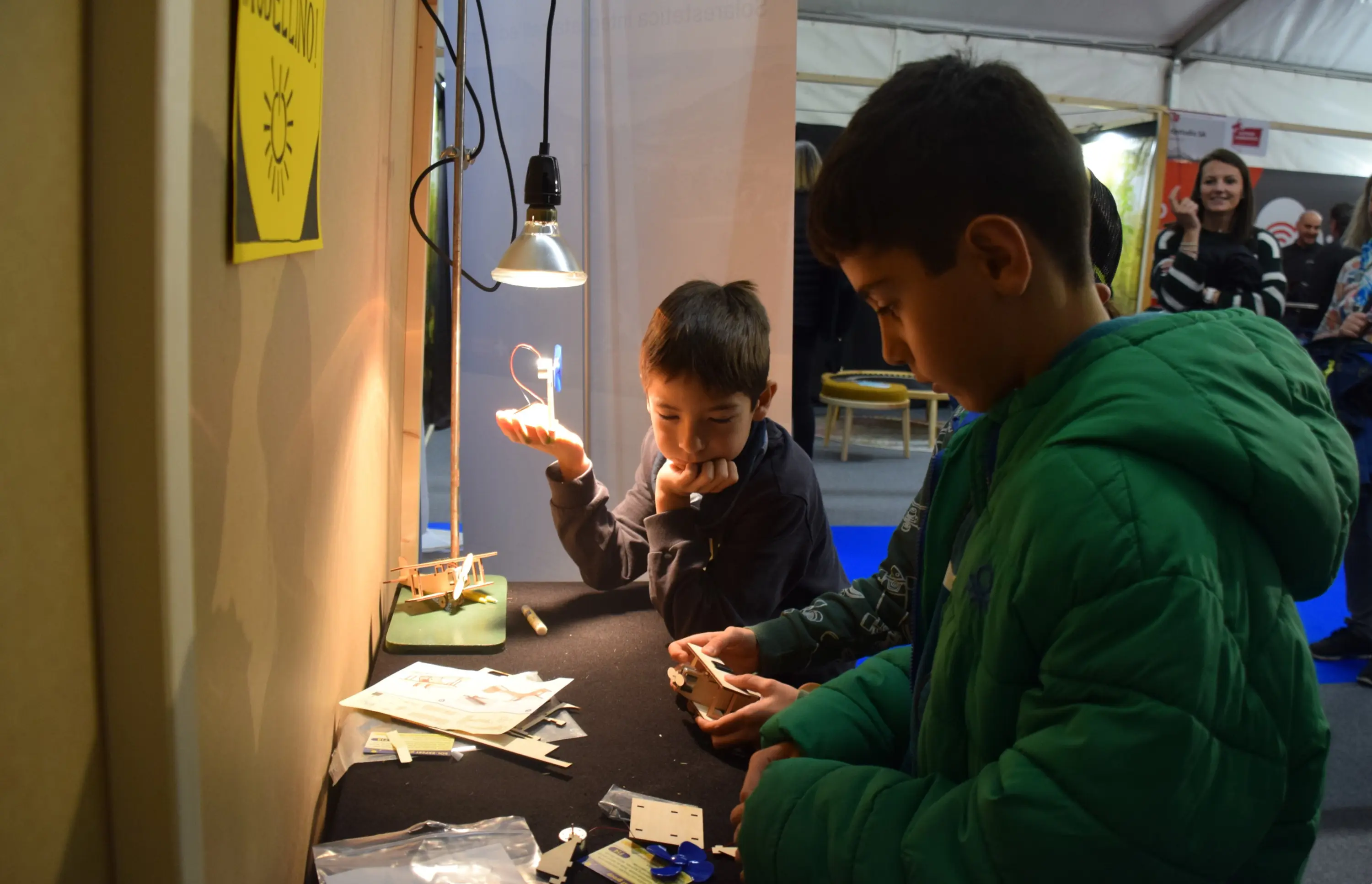 Due bambini stanno esplorando un tavolo da lavoro illuminato, pieno di materiali e strumenti. Un bambino indossa un giubbotto verde e osserva attentamente, mentre l'altro sembra pensieroso, con la mano al mento. Sul tavolo ci sono diversi oggetti e un'applique accesa. Sullo sfondo, si notano altre persone in un