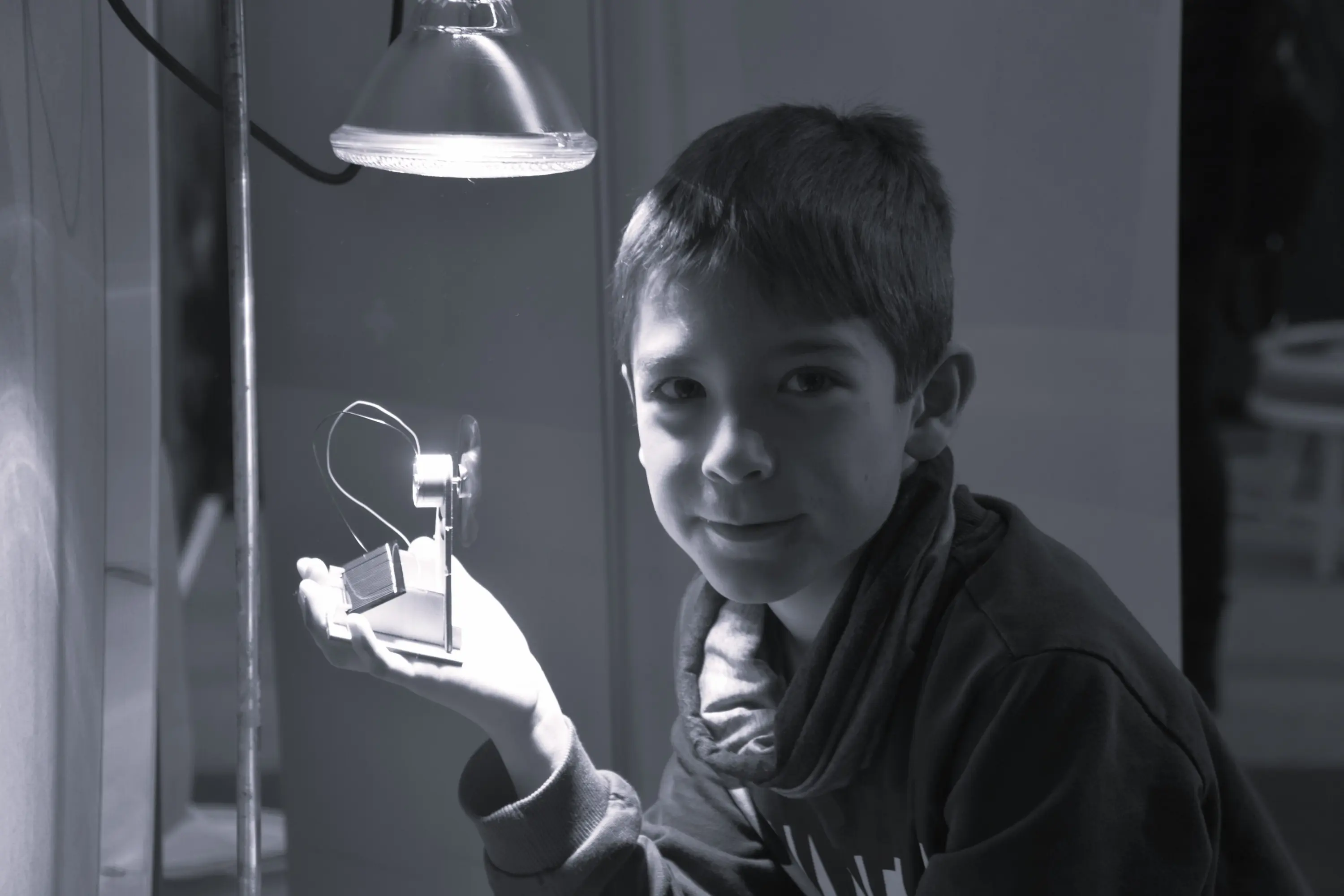 Ragazzo sorride mentre tiene un piccolo modello di un generatore elettrico, illuminato da una lampada. Il contrasto tra la luce e le ombre crea un'atmosfera interessante.