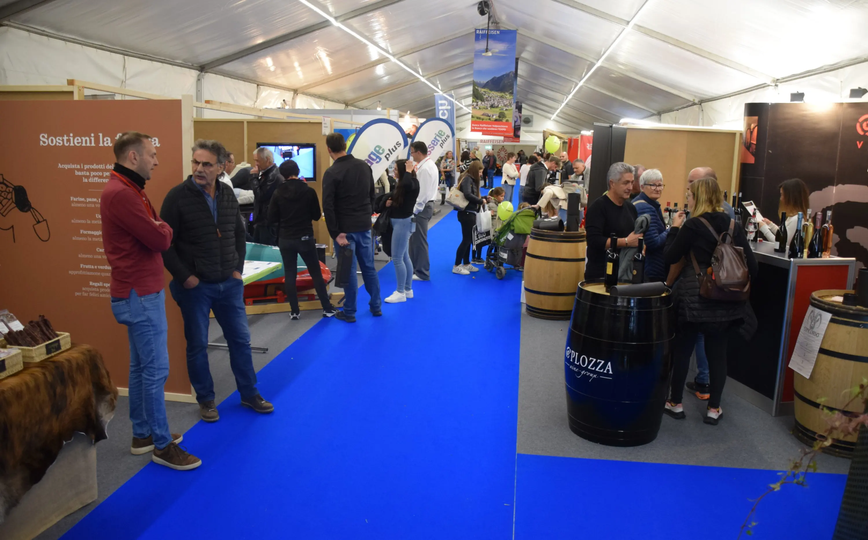 Una fiera affollata all'interno di un tendone. Persone si muovono tra casse di vino e stand, con un pavimento blu. Alcuni visitatori degustano prodotti, mentre altri osservano le esposizioni.