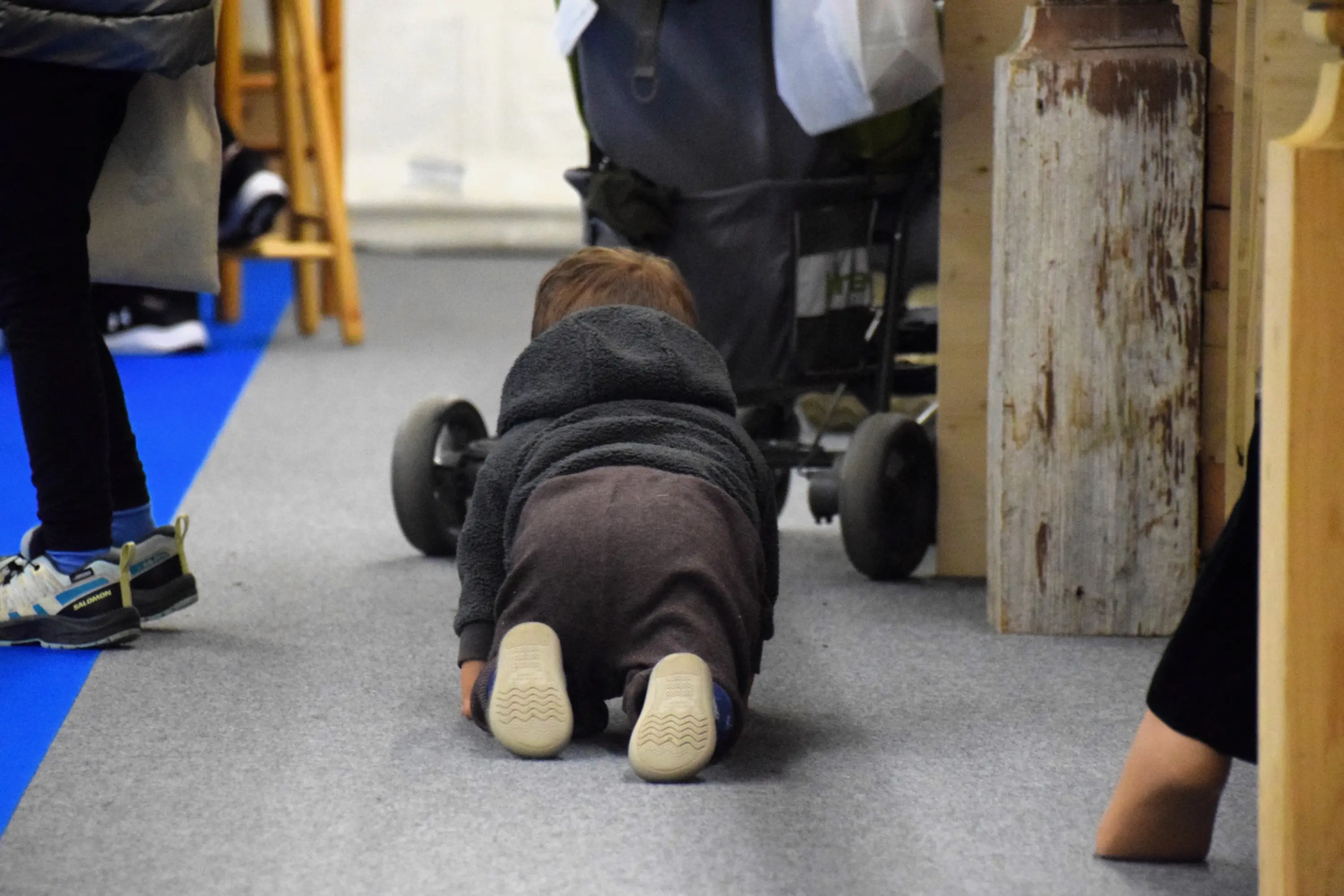 Un bambino str crawla sul pavimento in una zona commerciale, con un passeggino vicino. Indossa un maglione scuro e pantaloni marroni.