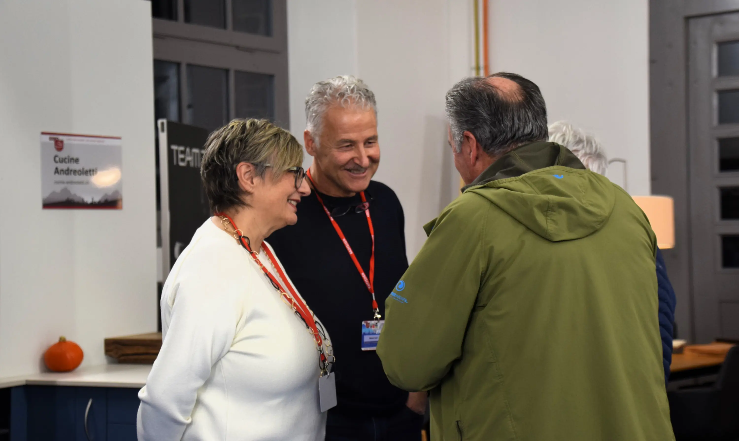 Tre persone conversano in un ambiente interno. Due uomini e una donna sorridono, mentre uno dei due uomini indossa una giacca verde. Sulla parete si intravede un cartello con il nome "Cucine Andreetti".