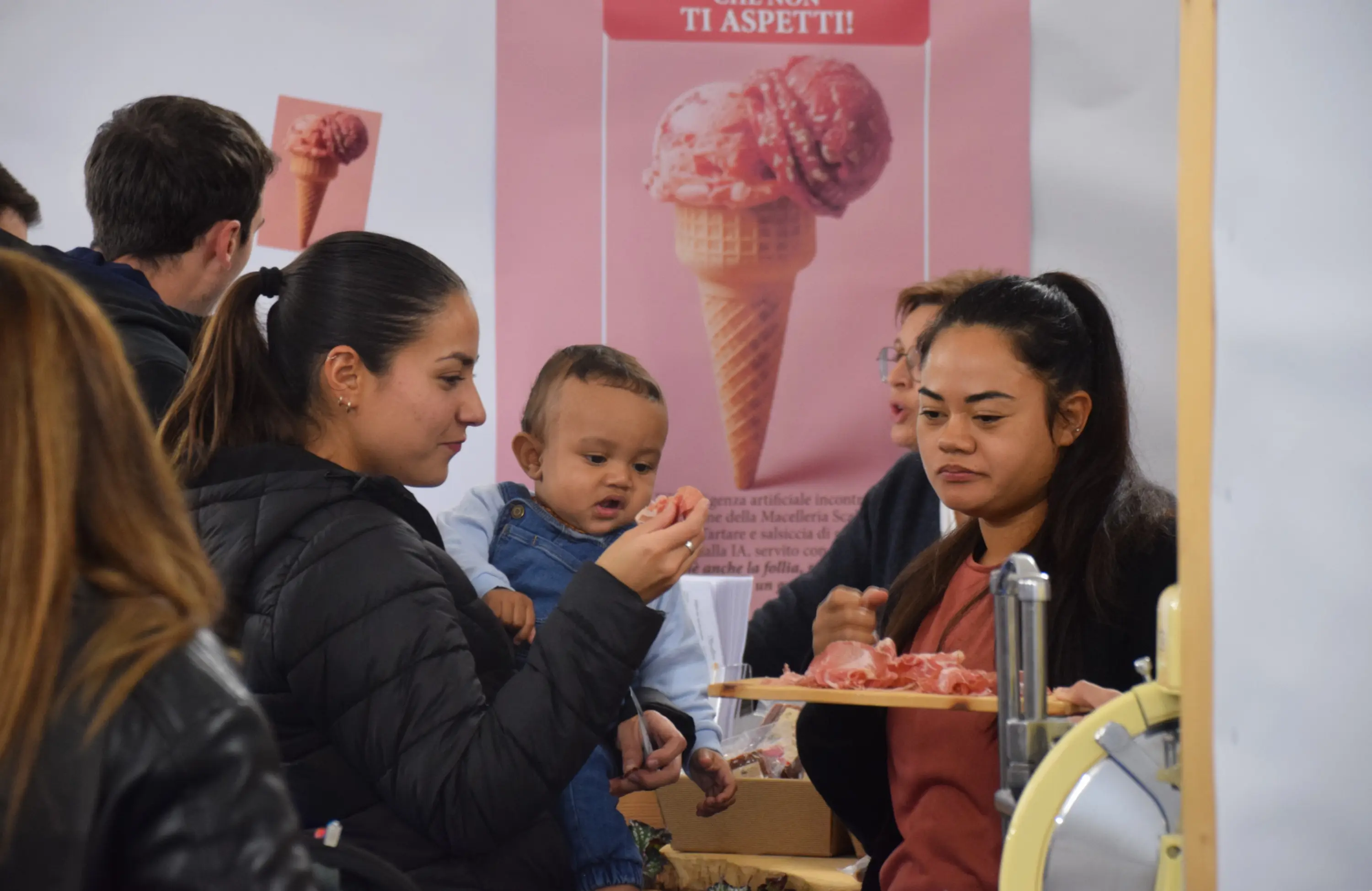 Due donne, una con un bambino in braccio, assaporano un campione di gelato, mentre altre persone si trovano sullo sfondo. Un cartellone con un cono gelato si intravede dietro di loro.