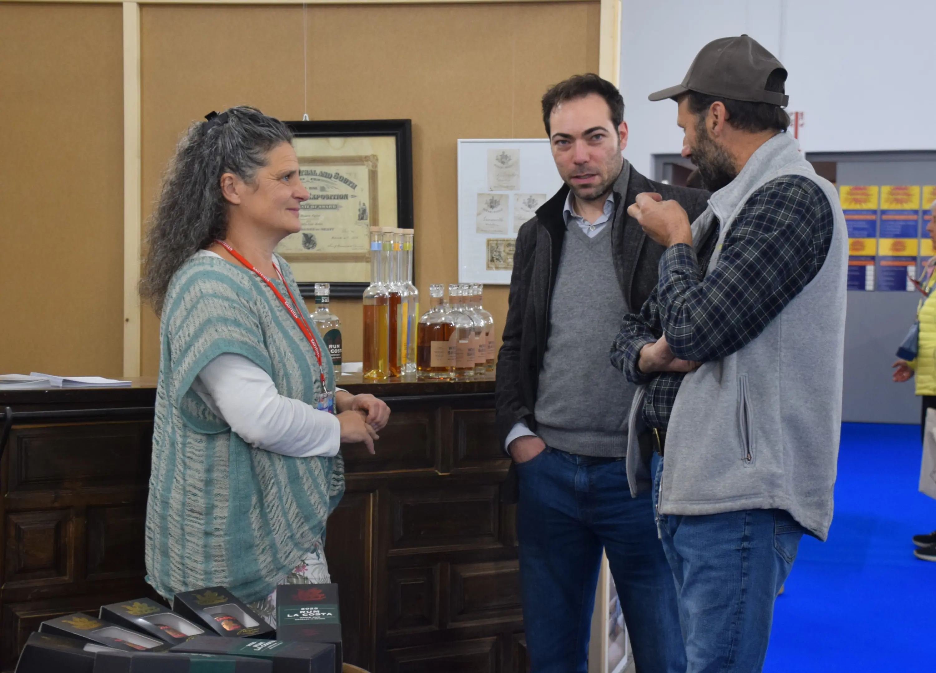 Un uomo e una donna conversano con un altro uomo in un ambiente di fiera. La donna, con capelli ricci e grigi, indossa un maglione bianco e una collana. Gli uomini, uno con giacca grigia e l'altro con cappellino e camicia a quadri, sembrano interessati ai prodotti esposti. Uno sfondo con un