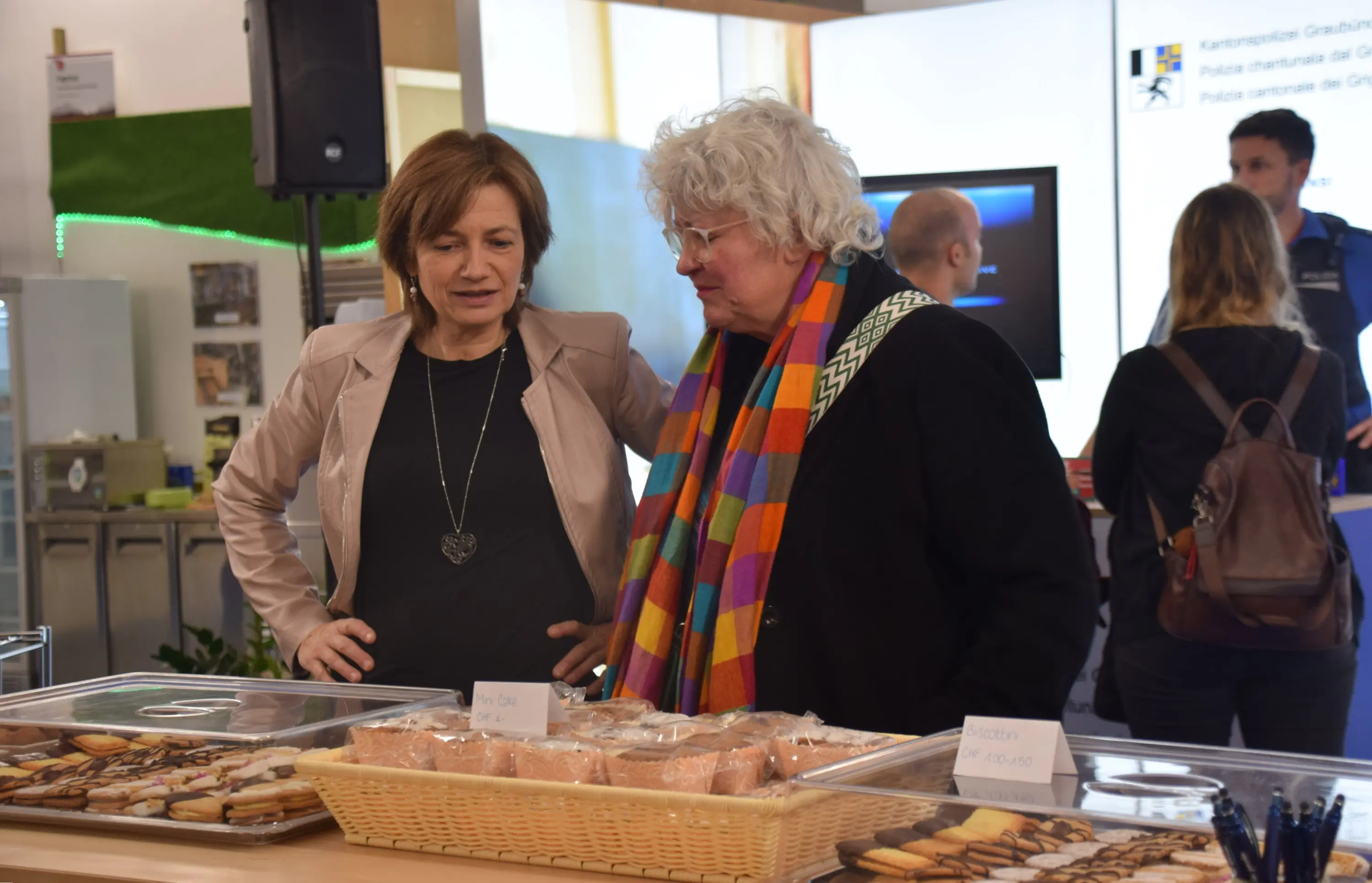 Due donne osservano un espositore di dolci, con biscotti e dessert, mentre discutono tra loro. Sullo sfondo ci sono altre persone e un display informativo.
