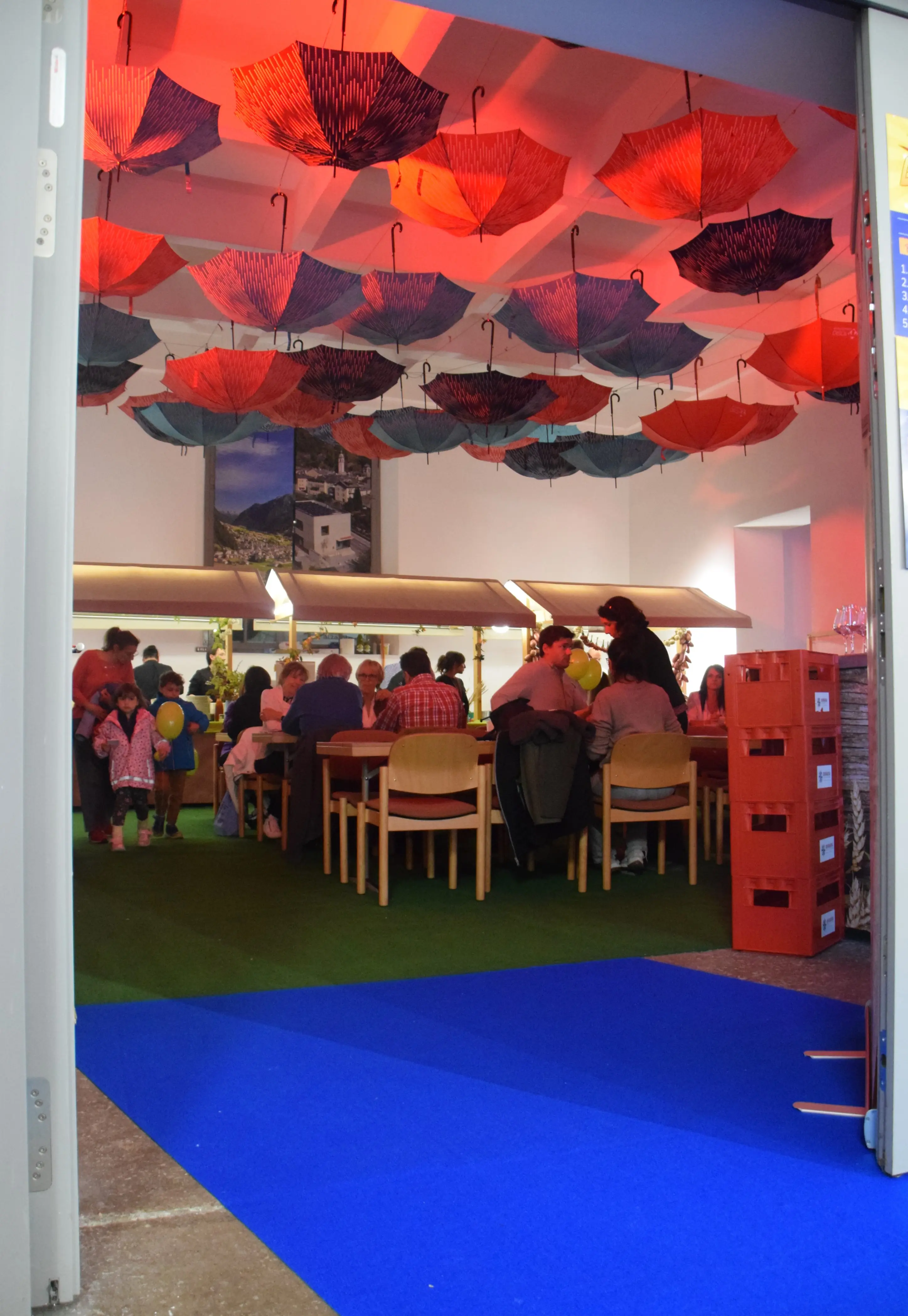 Una sala con un soffitto decorato con ombrelli rossi e blu. Diverse persone siedono a tavoli. L'ambiente è colorato e accogliente, con un tappeto verde e un ingresso blu.