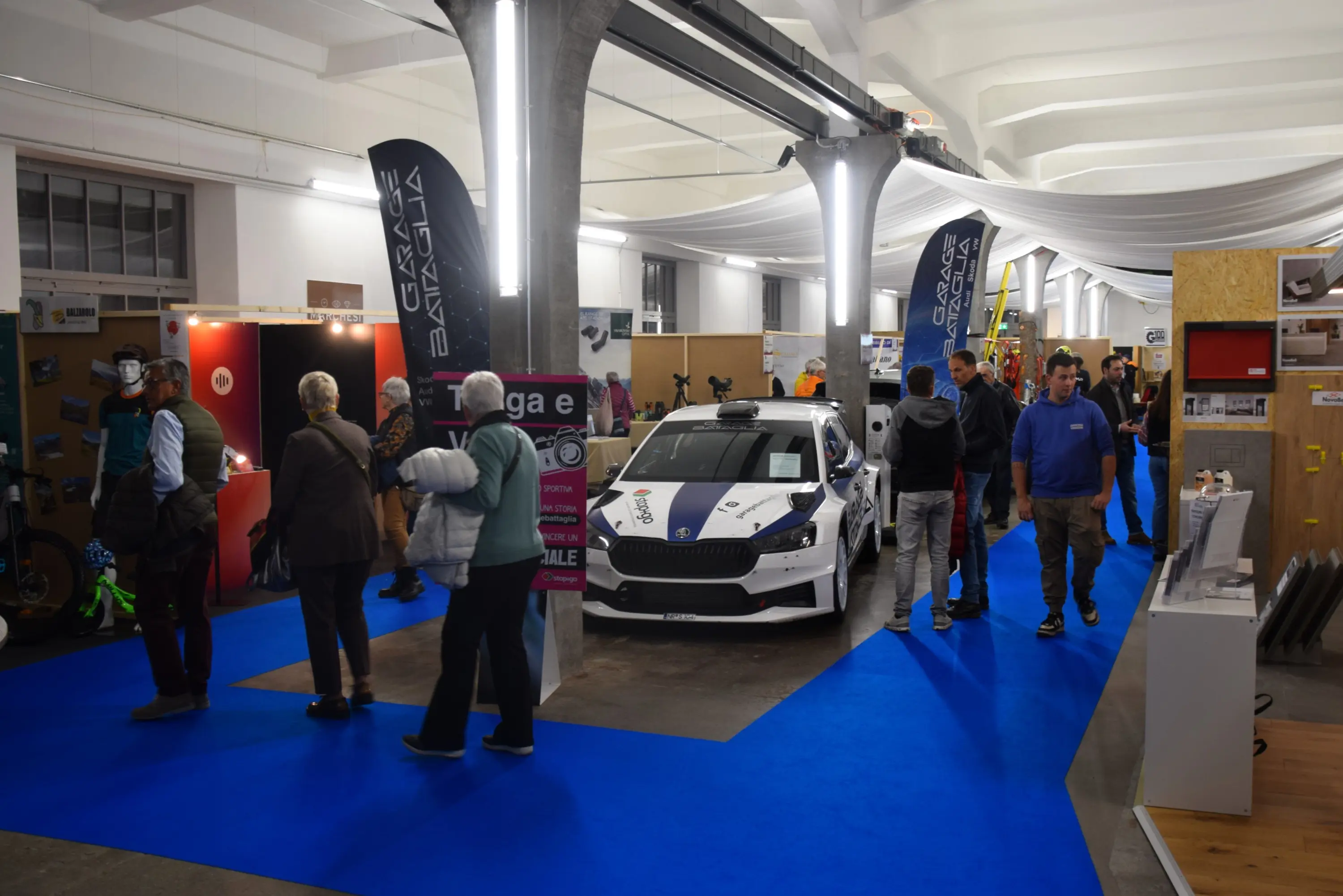 Un evento espositivo con persone che passeggiano tra vari stand. In primo piano c'è un'auto da corsa su una superficie blu, circondata da poster e attrezzature sportive. Sullo sfondo, visitatori osservano diversi prodotti e attrezzature.