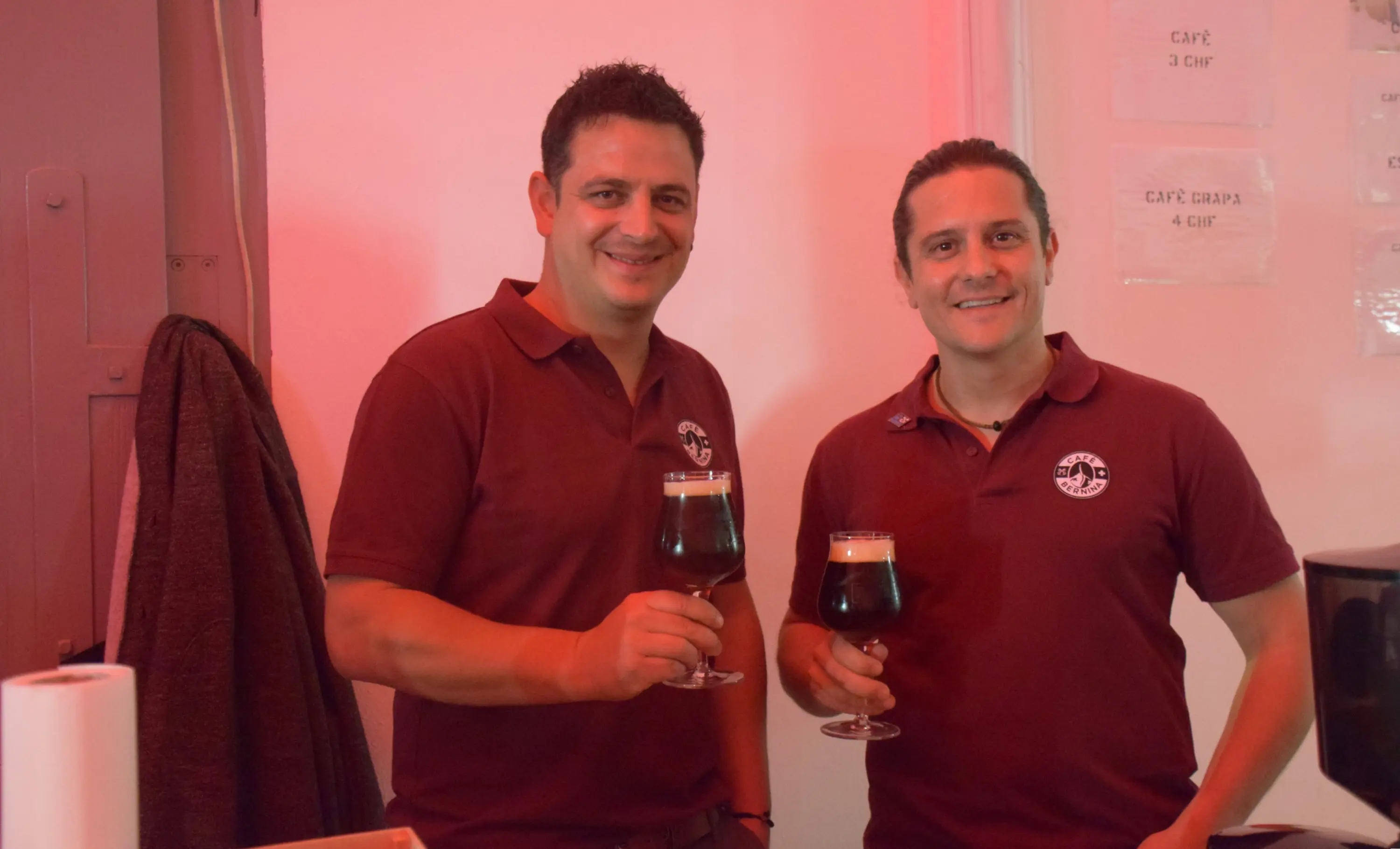 Due uomini sorridenti indossano magliette bordeaux e tengono in mano bicchieri di birra scura. Sullo sfondo, una parete con luce rossa e alcune informazioni scritte.