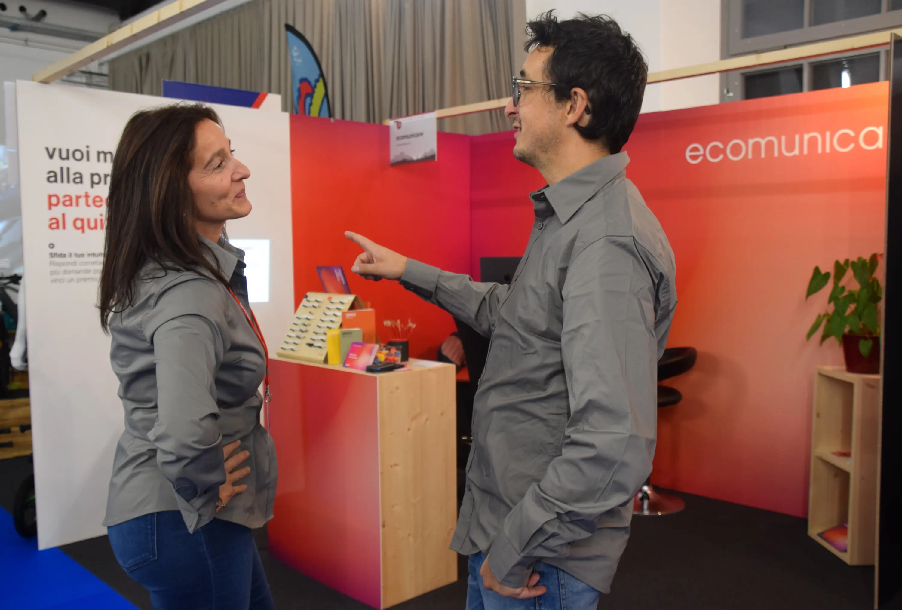 Due persone sorridenti interagiscono a uno stand espositivo. Entrambi indossano camicie grigie e sembrano impegnati in una conversazione amichevole. Sullo sfondo, si nota un'area con un design colorato e uno slogan in italiano.
