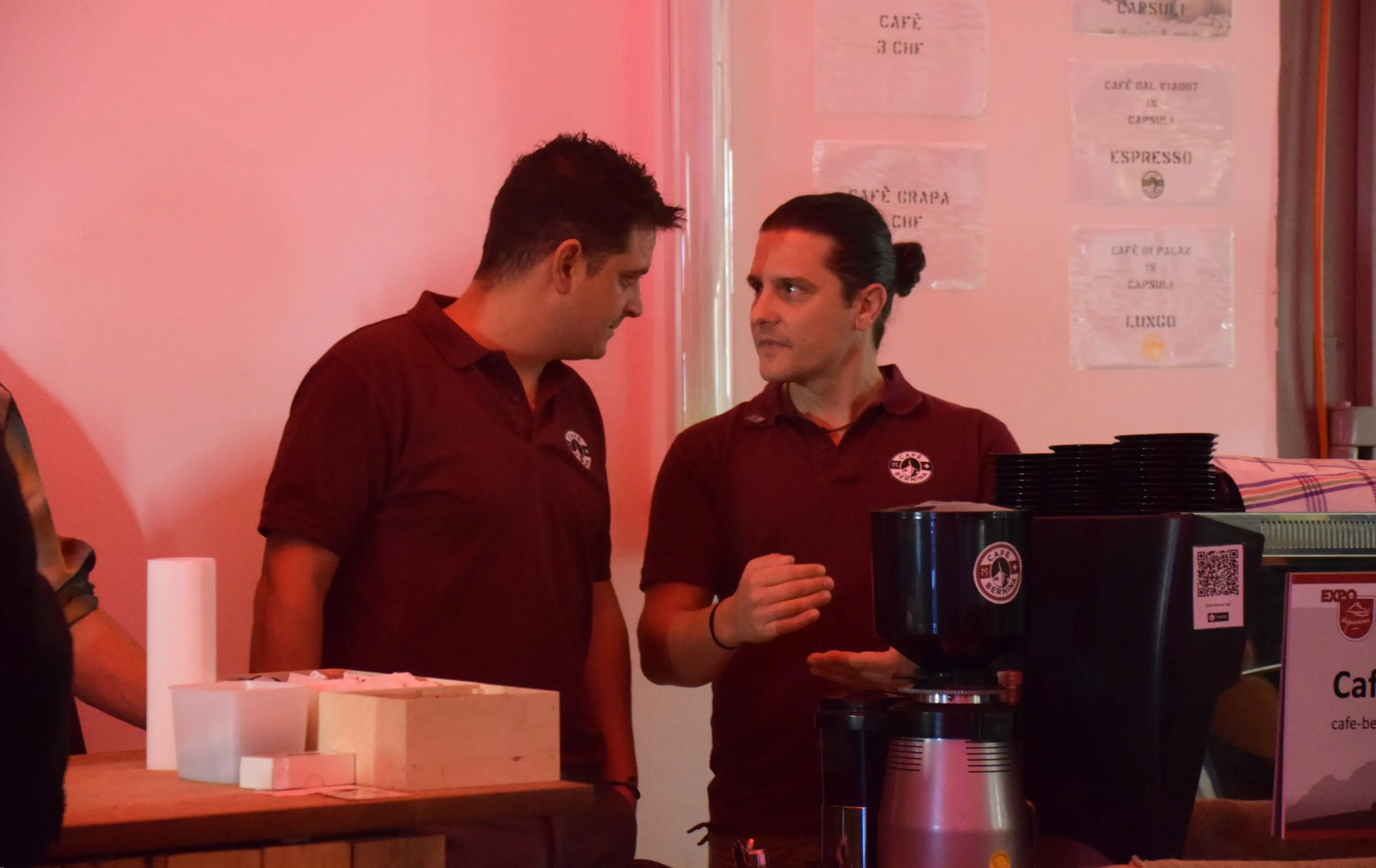 Due uomini in maglietta rossa discutono dietro un bancone di caffè. Sullo sfondo, si possono vedere vari cartelli e strumenti per il caffè.