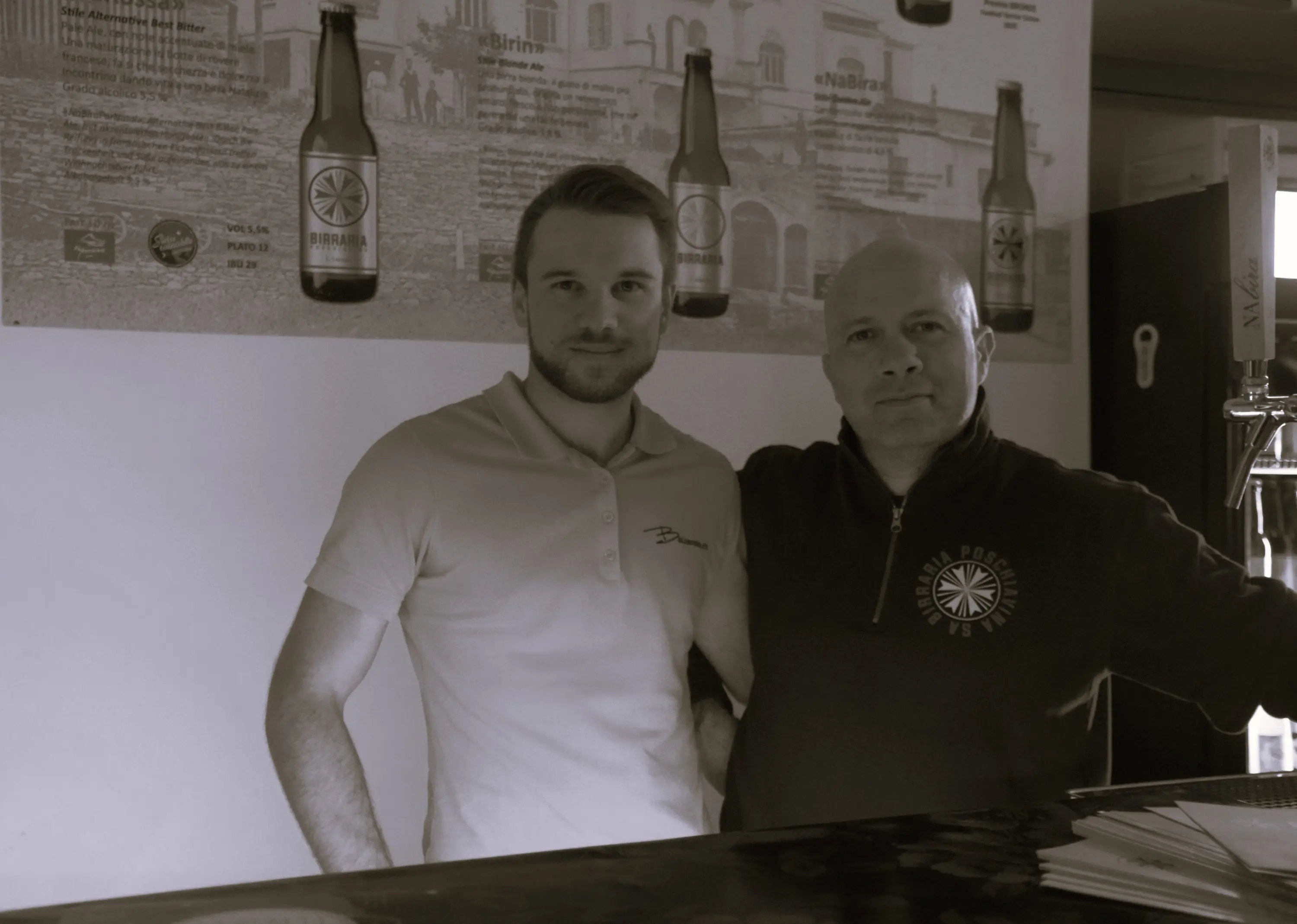 Due uomini sorridenti in un bar, uno con una maglietta chiara e l'altro con un pullover scuro. Sullo sfondo, un poster che pubblicizza birre.