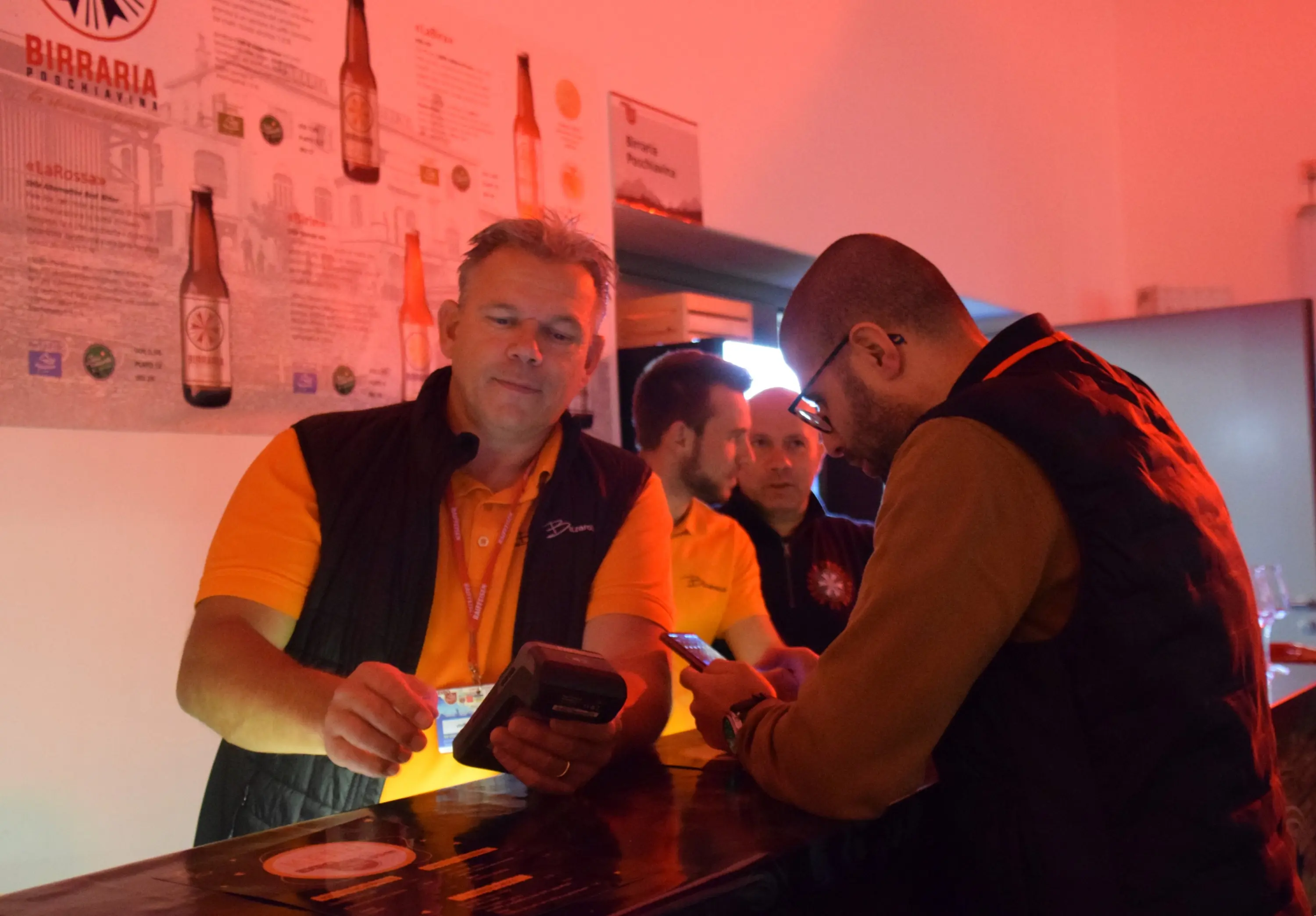 Un barista sorride mentre usa un registratore di cassa per servire un cliente. Sullo sfondo, altri membri dello staff sono visibili. L'illuminazione è rossa, creando un'atmosfera vivace.