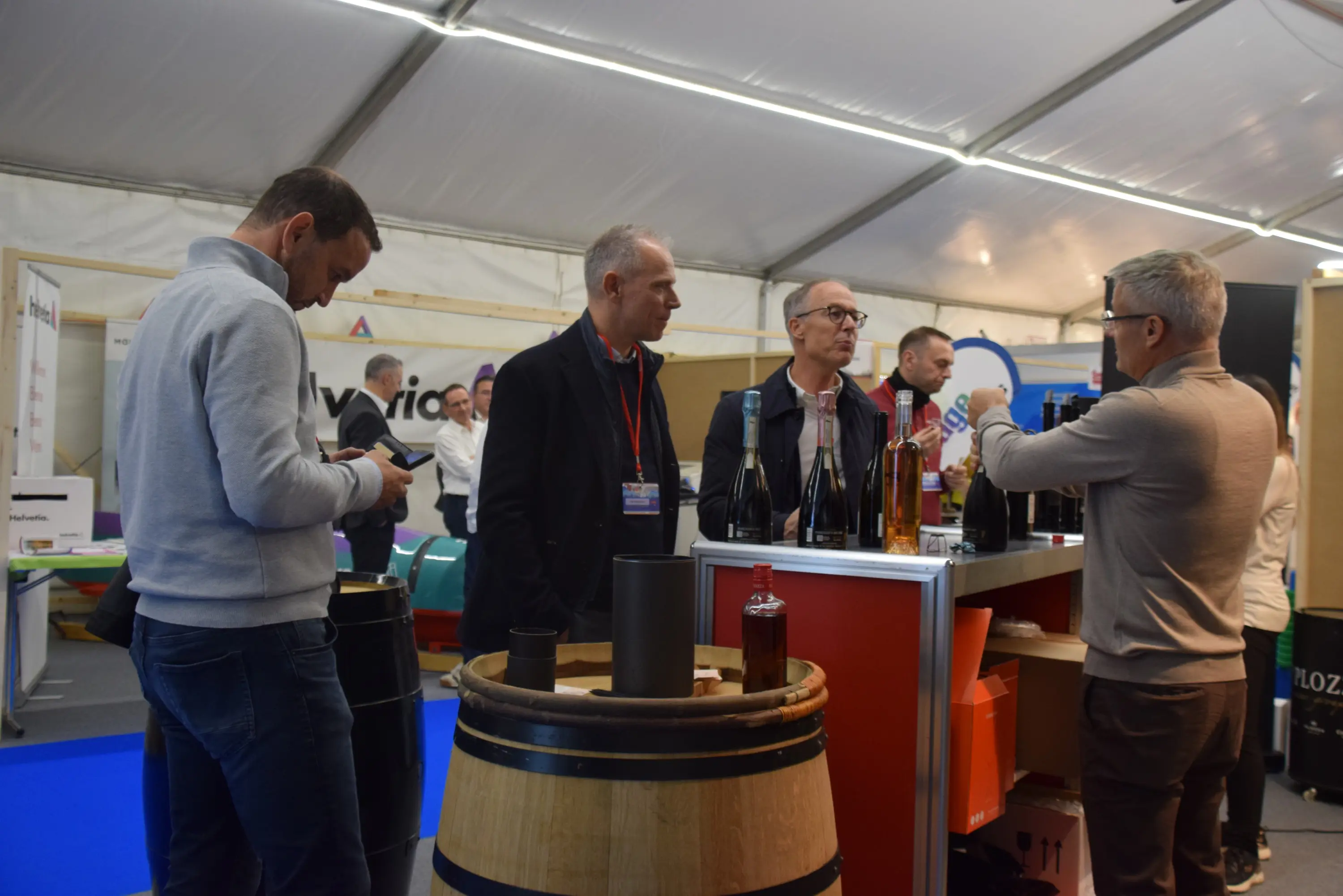 Un gruppo di uomini in un'area espositiva, mentre discutono davanti a un bancone con bottiglie di vino. Alcuni osservano le etichette, altri sono impegnati con i telefoni. Ambientazione all'interno di una tenda con una decorazione commerciale.
