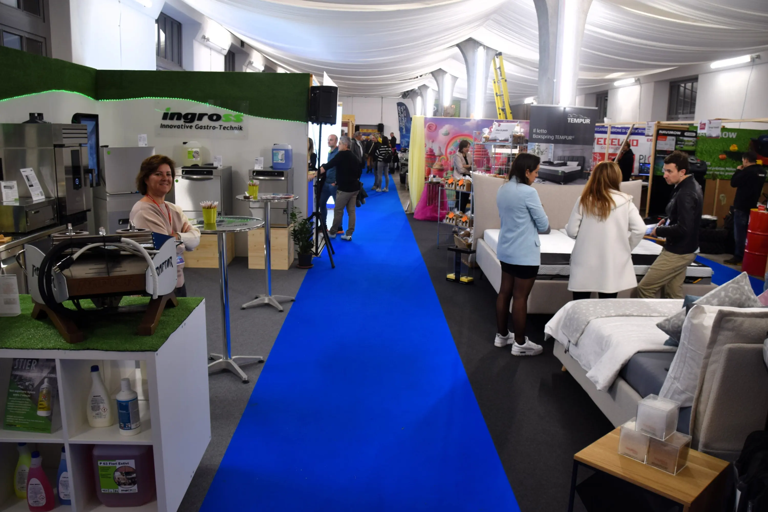 Fiera con un corridoio centrale blu, espositori di prodotti per la casa e persone che interagiscono. Si vede una donna sorridente accentata da piante, mentre altri visitatori si fermano a esaminare varie attrezzature e mobili.