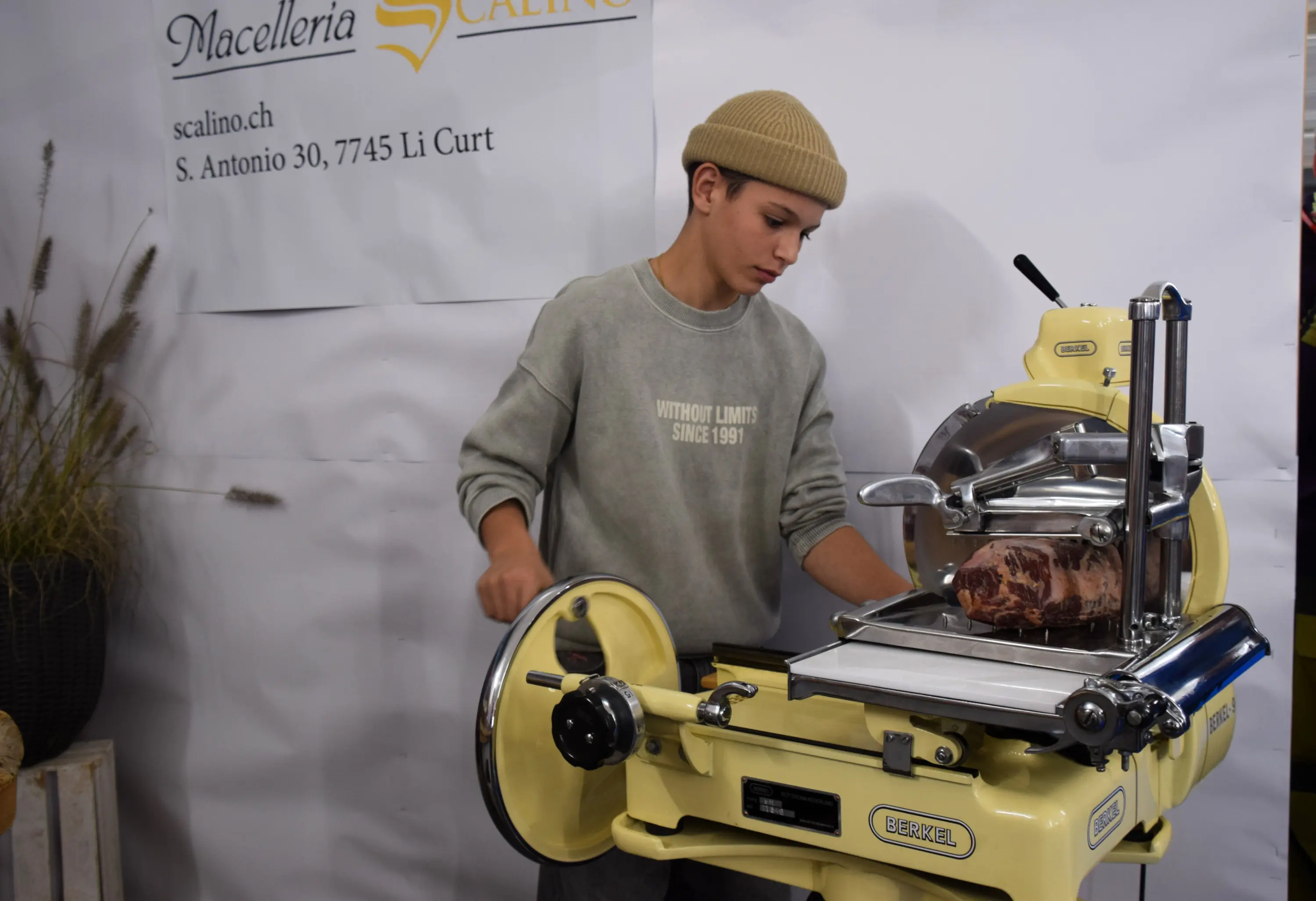 Un giovane sta utilizzando un'affettatrice vintage di colore giallo per tagliare un pezzo di carne. Indossa un maglione grigio e un berretto. Sullo sfondo si intravede un'insegna con il nome "Macelleria Scalino".