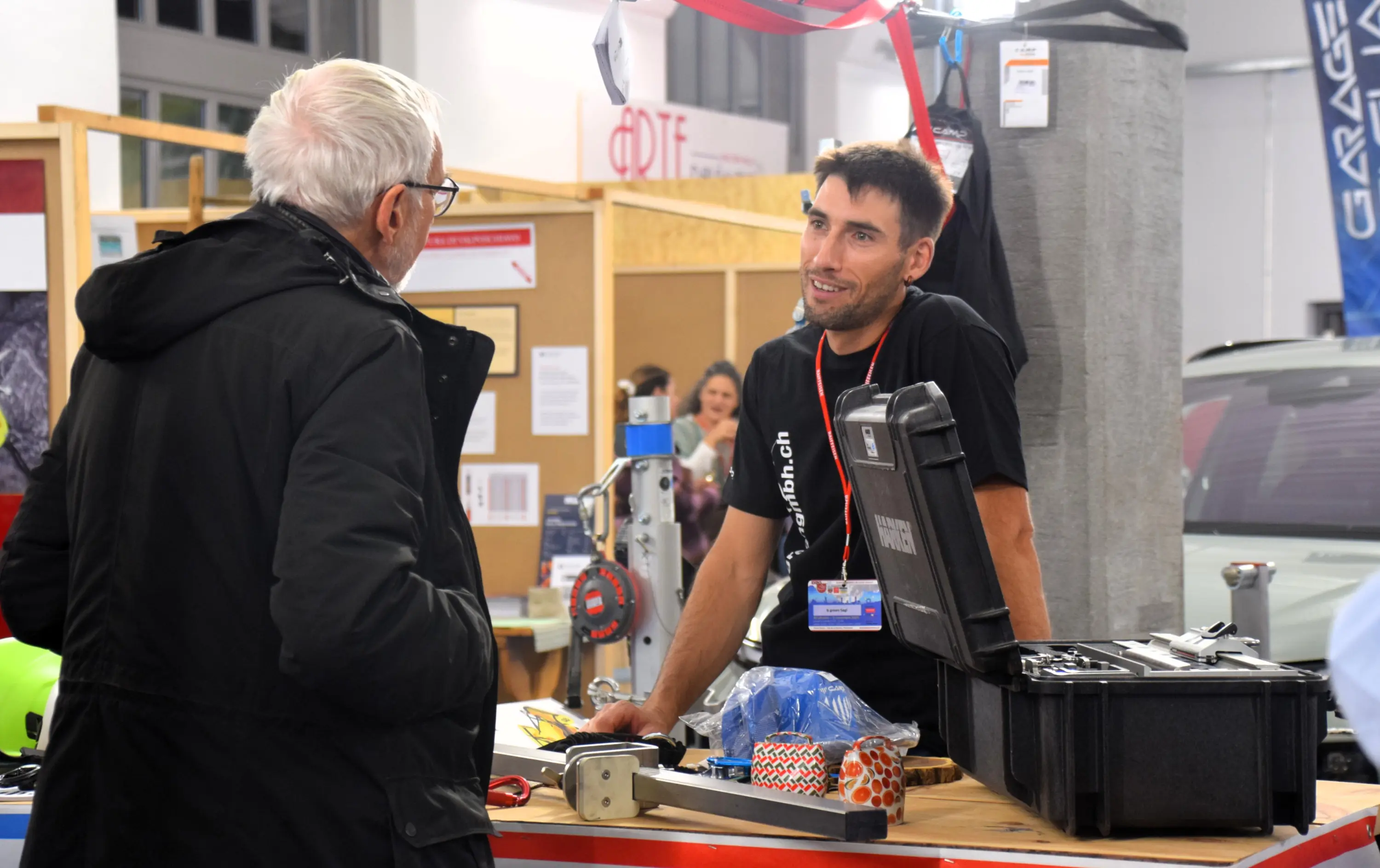 Due uomini stanno conversando a un evento, con attrezzature da lavoro e strumenti su un tavolo. L'uomo con i capelli chiari indossa occhiali e un cappotto nero, mentre l'altro indossa una maglietta nera. Si intravede un fondale con altre persone e attrezzature.