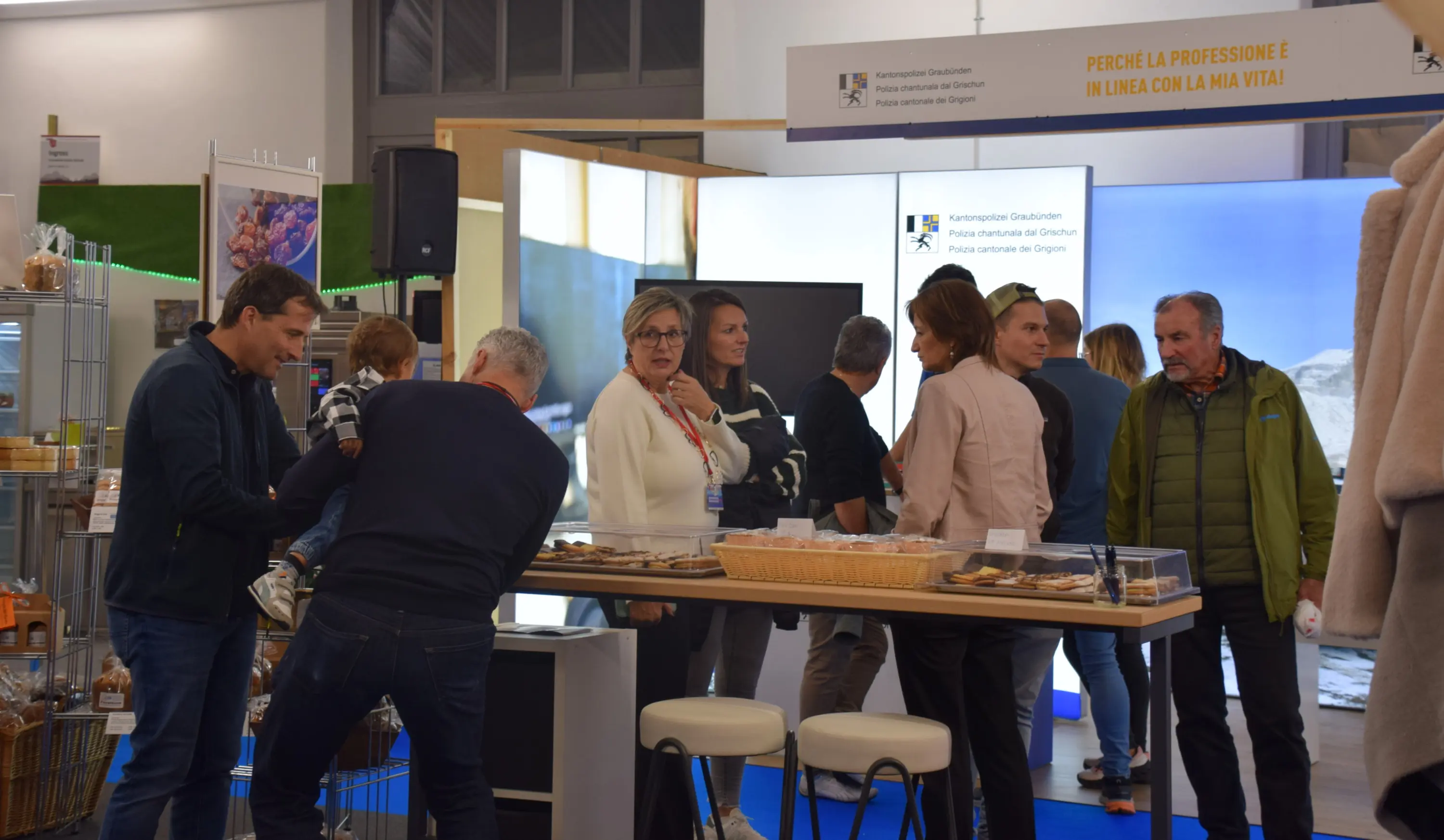 Un gruppo di persone interagisce all'interno di un evento espositivo. Alcuni visitatori sono intenti a guardare e degustare prodotti alimentari, mentre altri conversano tra loro. Sullo sfondo si scorge uno stand con schermi e prodotti in mostra.