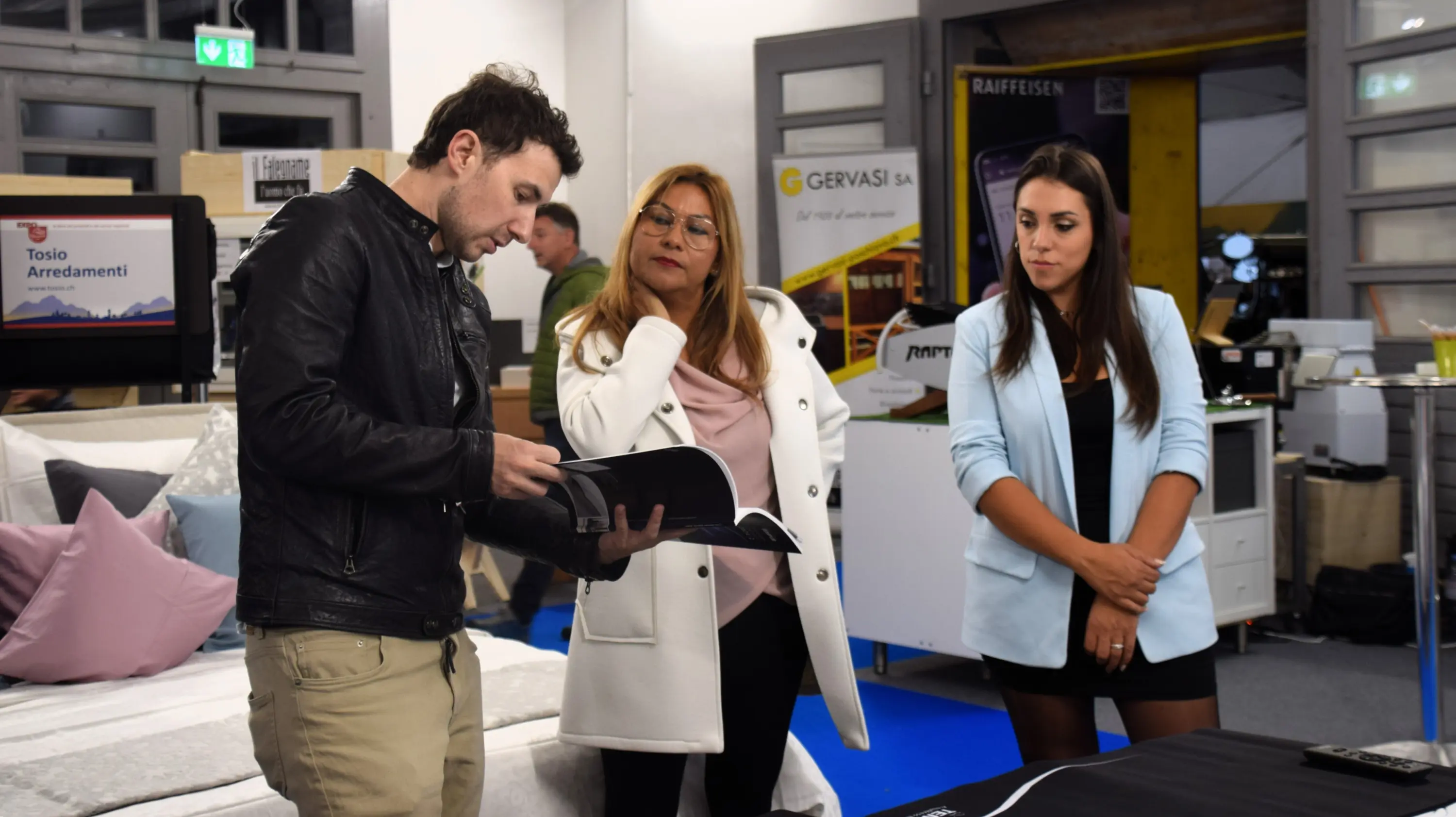 Tre persone in una sala espositiva. Un uomo in giacca nera sfoglia un catalogo, mentre due donne lo osservano attentamente. Una donna ha i capelli lunghi e castani e indossa una maglietta bianca, l'altra ha capelli biondi e indossa un abito elegante. Sullo sfondo ci sono espositori di arredamento.