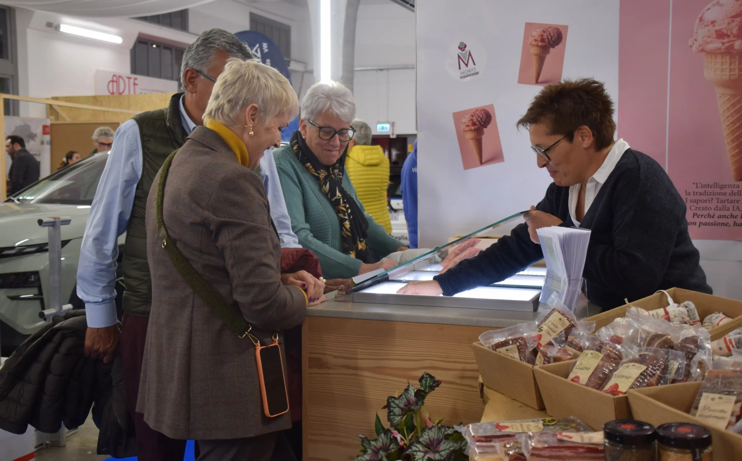 Persone interessate in un mercato di prodotti alimentari, mentre osservano una vetrina di gelati. Una donna dietro il bancone serve i clienti, che discutono tra loro. Sullo sfondo, si intravedono altri visitatori e stand.