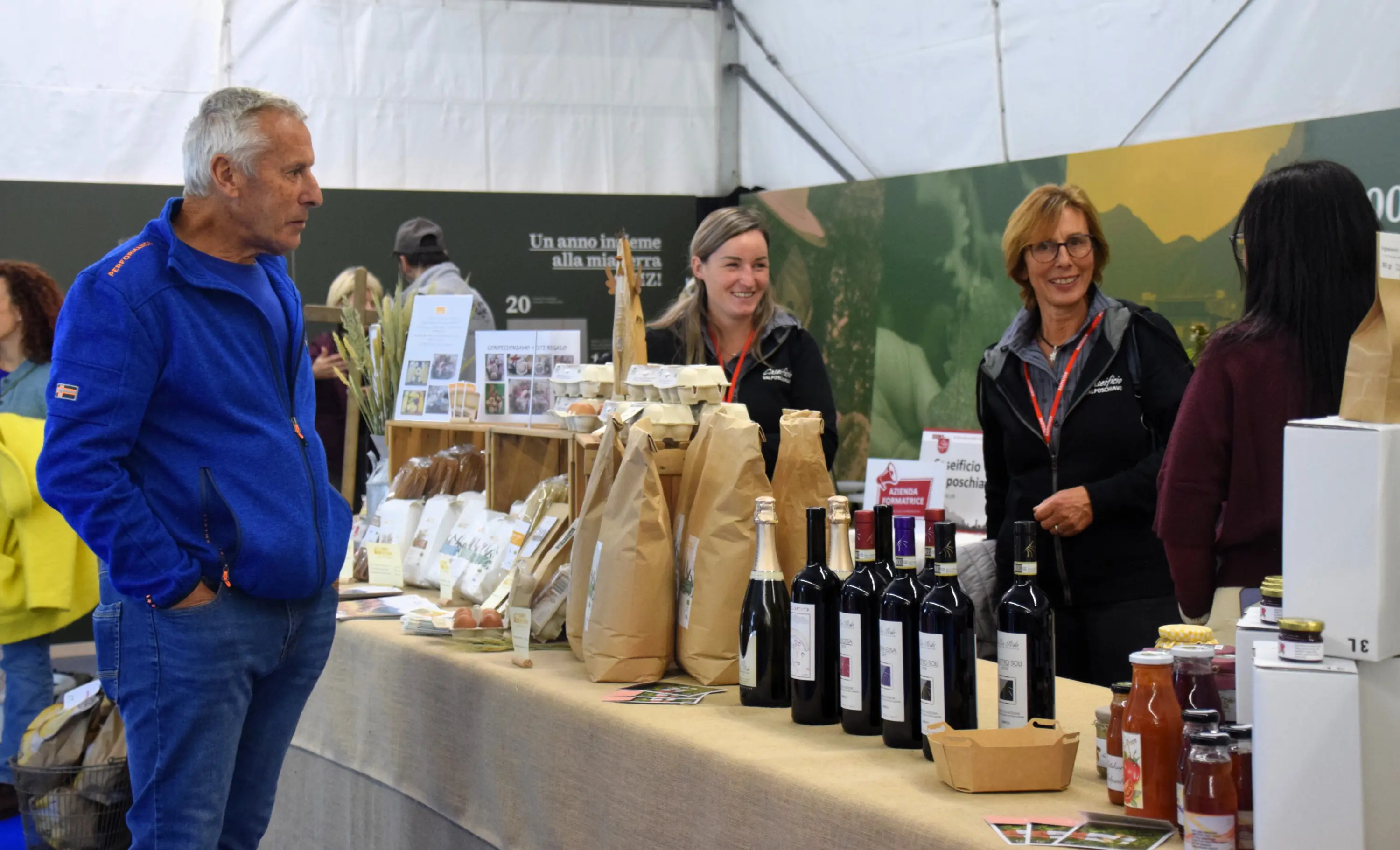 Un gruppo di persone interagisce a un bancone con prodotti locali, tra cui vini e alimenti confezionati. Sullo sfondo, un cartellone con informazioni sulla promozione dei prodotti tipici.