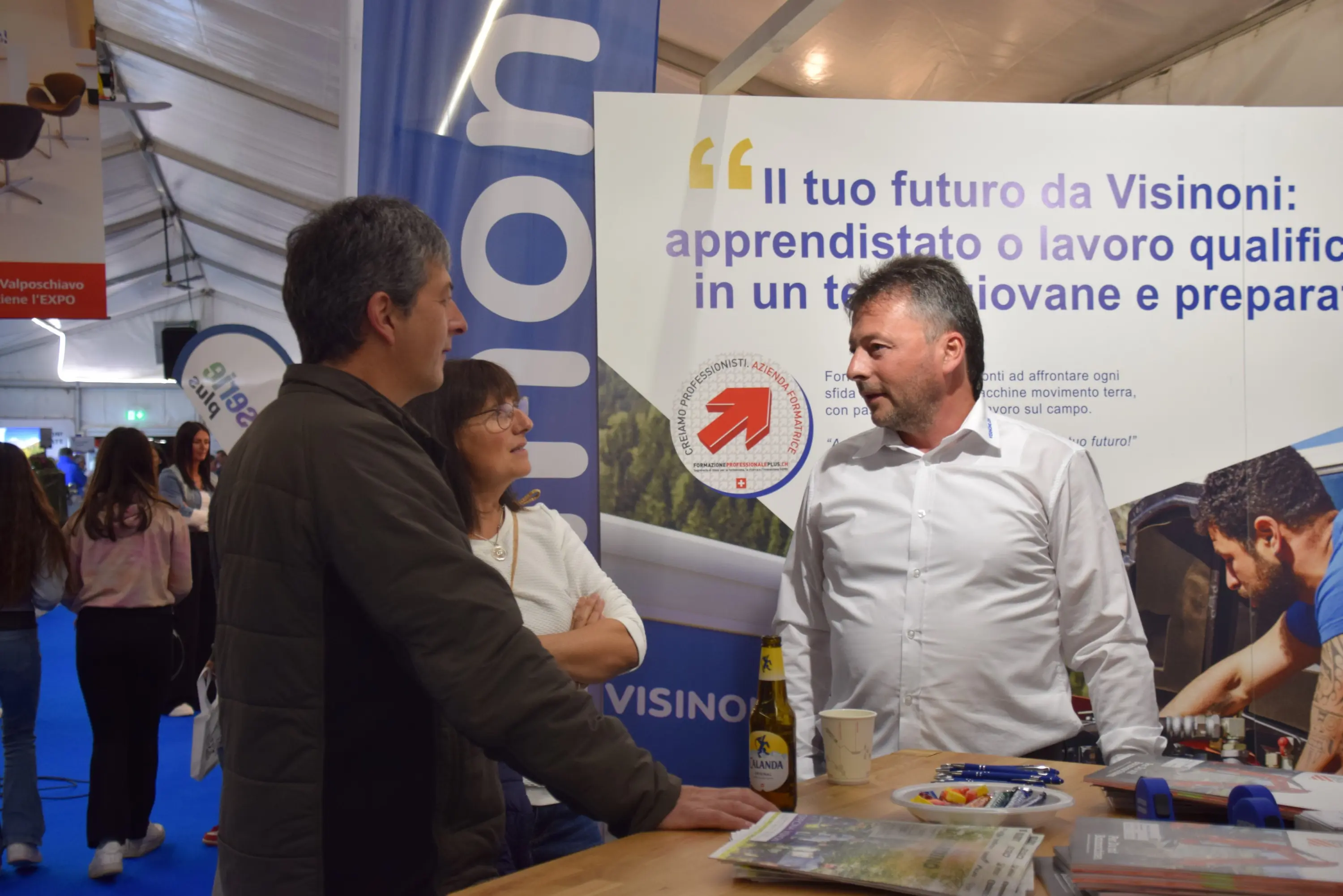 Tre persone discutono a uno stand di Visiononi durante un evento, con un banner che promuove opportunità di apprendimento e lavoro.