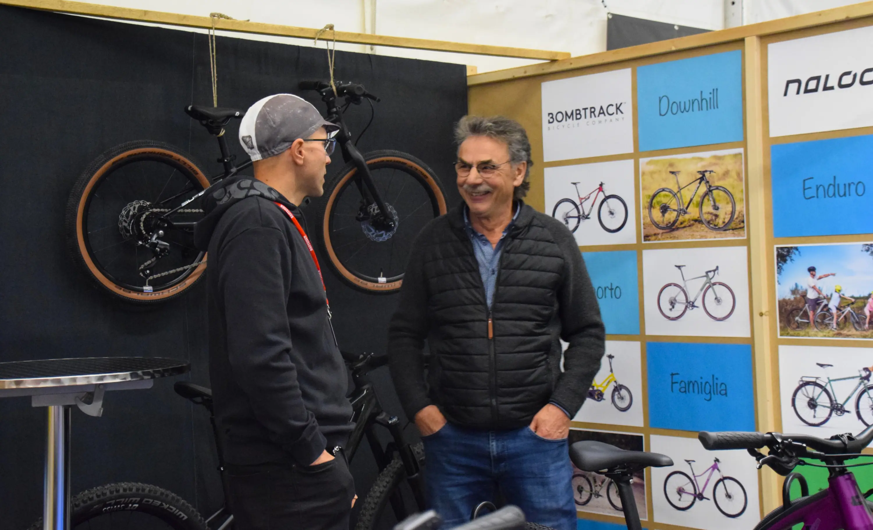 Due uomini sorridenti conversano in un negozio di biciclette, circondati da vari modelli appesi al muro.