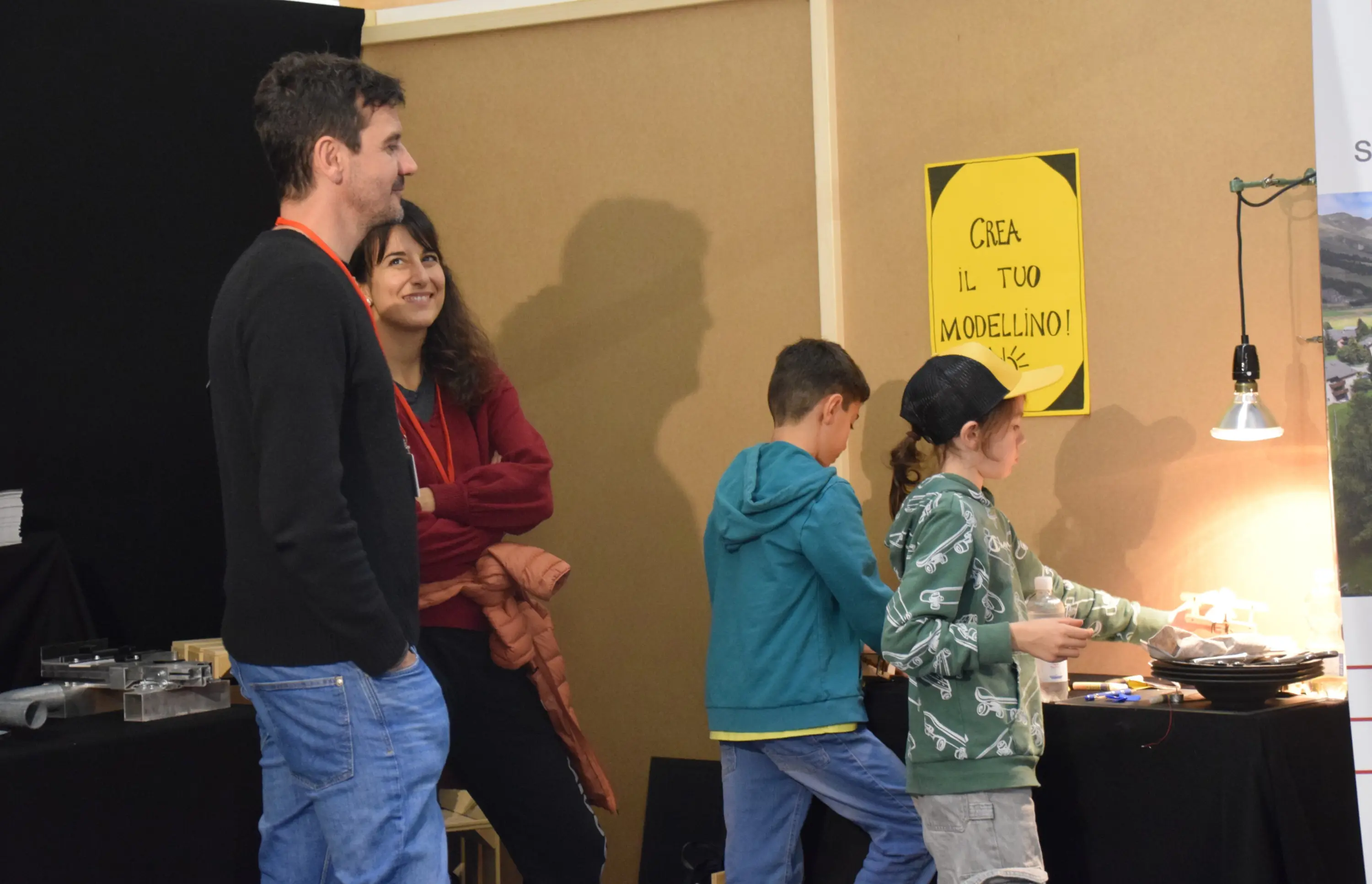 Un gruppo di persone in un laboratorio creativo. Due adulti osservano due bambini mentre lavorano su un progetto artistico. Uno dei bambini indossa un felpa verde e l'altro una maglietta blu. Sullo sfondo, un cartello giallo invita a "Creare il tuo modellino".