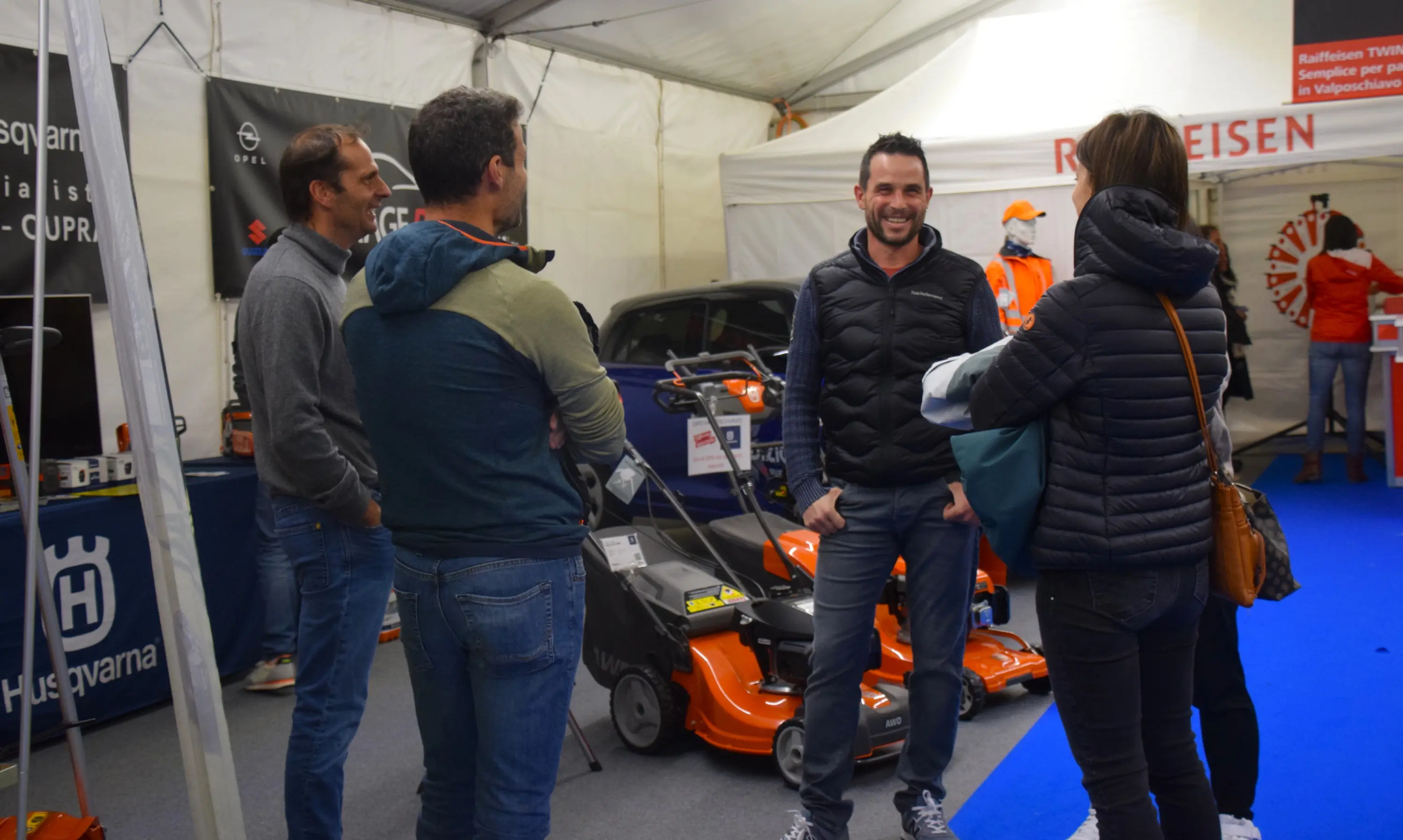 Quattro persone conversano in un'area espositiva all'interno di una tenda. Due uomini e due donne sono vestiti con giacche invernali. Sullo sfondo si intravede un'auto e attrezzature per il giardinaggio.