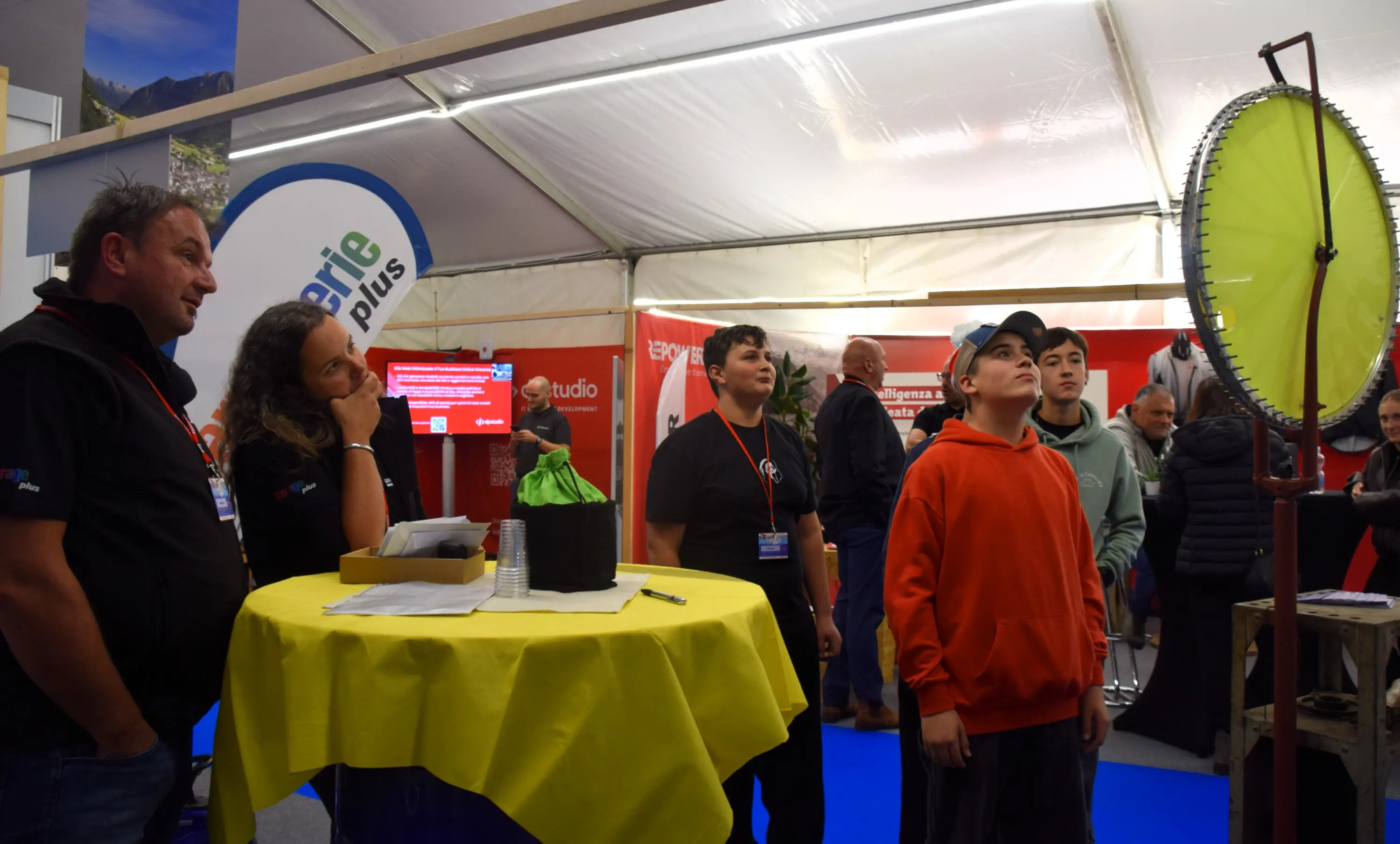 Un gruppo di persone osserva una ruota della fortuna all'interno di una fiera. Sono presenti due adulti e tre ragazzi, tutti concentrati sull'evento. Lo stand presenta banner e decorazioni colorate.