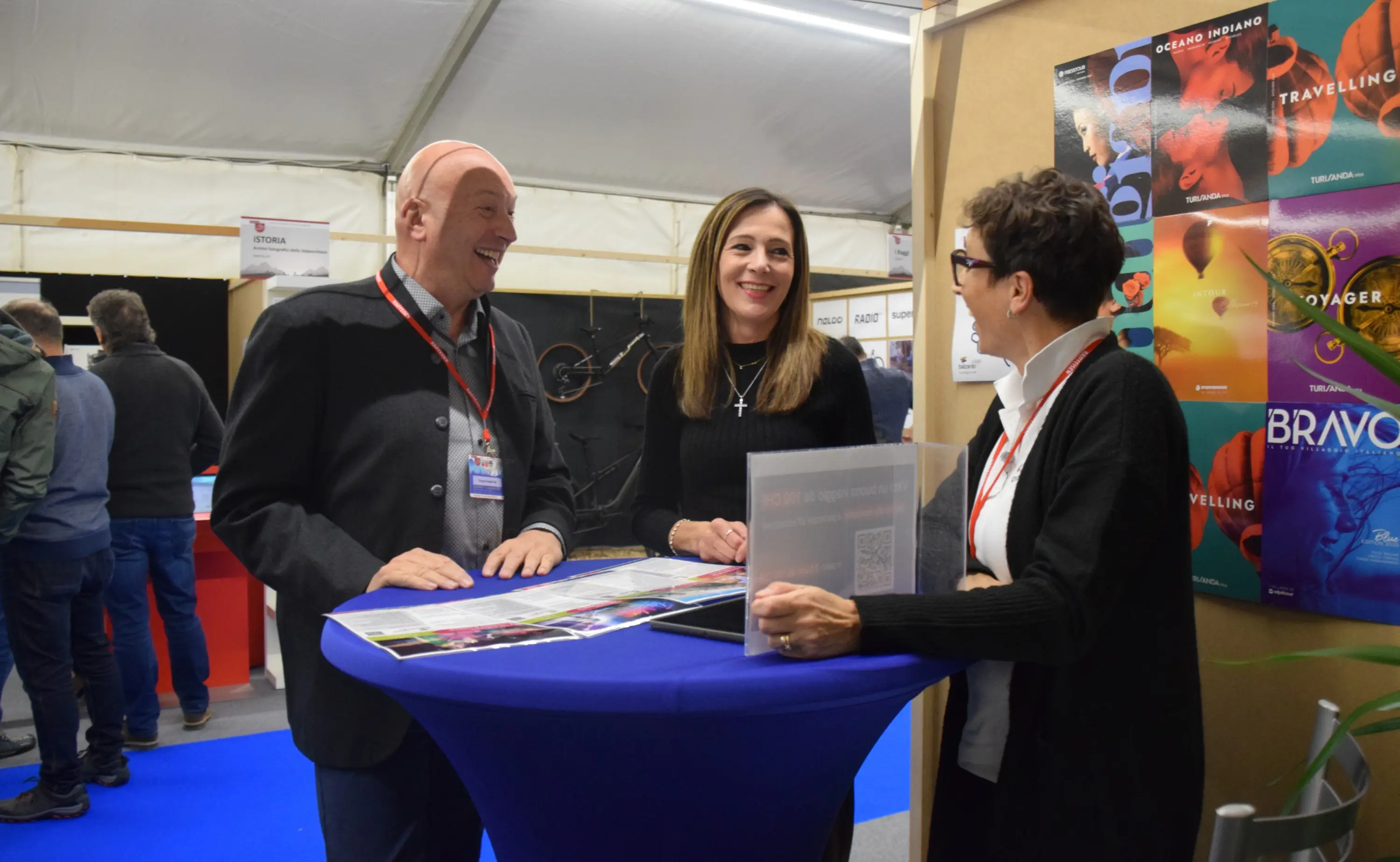 Tre persone sorridenti conversano attorno a un tavolo blu in un ambiente fieristico. Sono presenti materiali promozionali sul tavolo e poster colorati sullo sfondo.