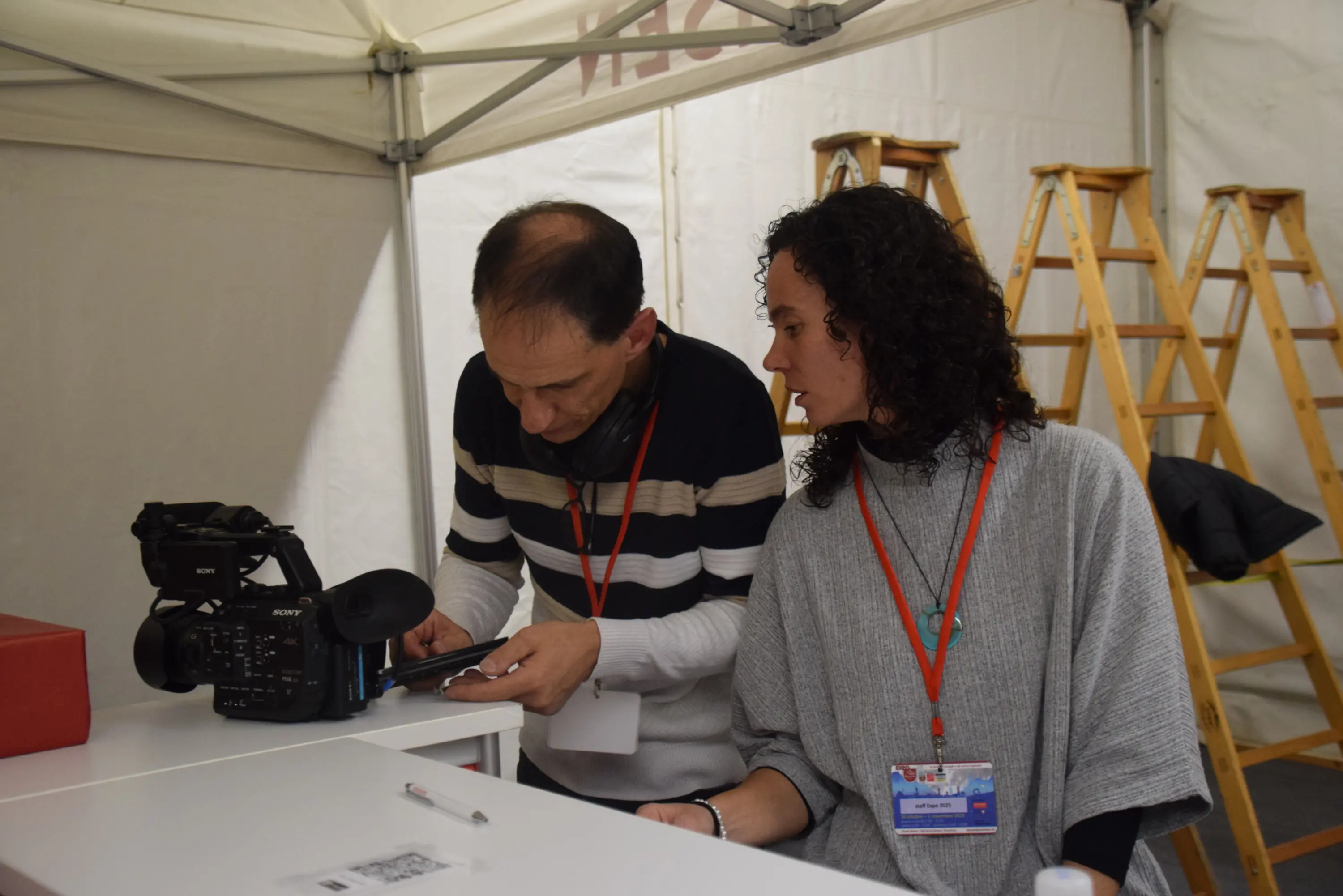 Due persone discutono davanti a una videocamera. Un uomo guarda attentamente un telefono, mentre una donna, con i capelli ricci e scuri, lo osserva. Entrambi indossano un pass per l'evento e si trovano in un ambiente all'interno di una tenda.