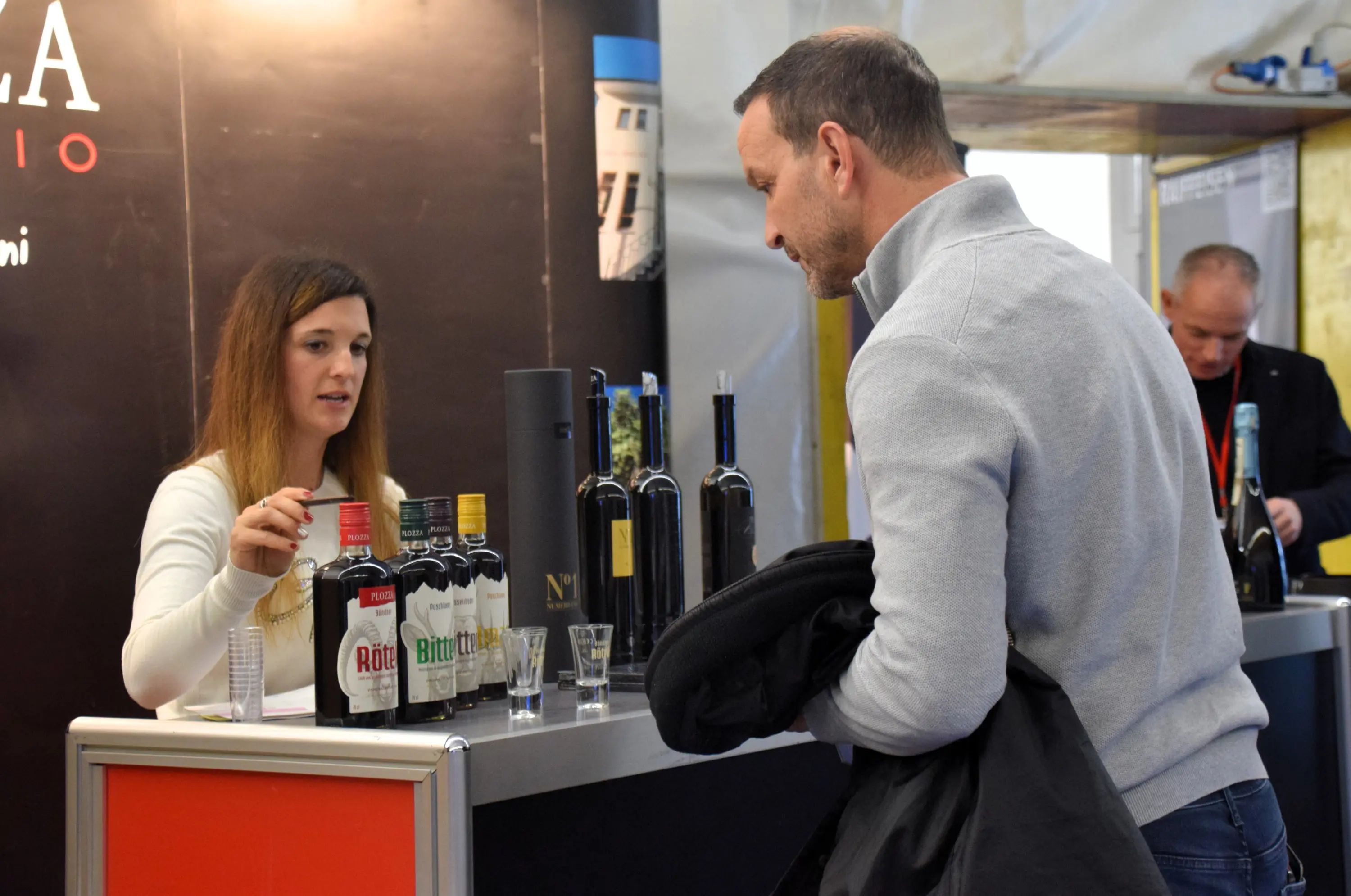 Una donna espone diverse bottiglie di vino a un bancone mentre un uomo ascolta attentamente. Sullo sfondo, altri visitatori interagiscono in un evento di degustazione.