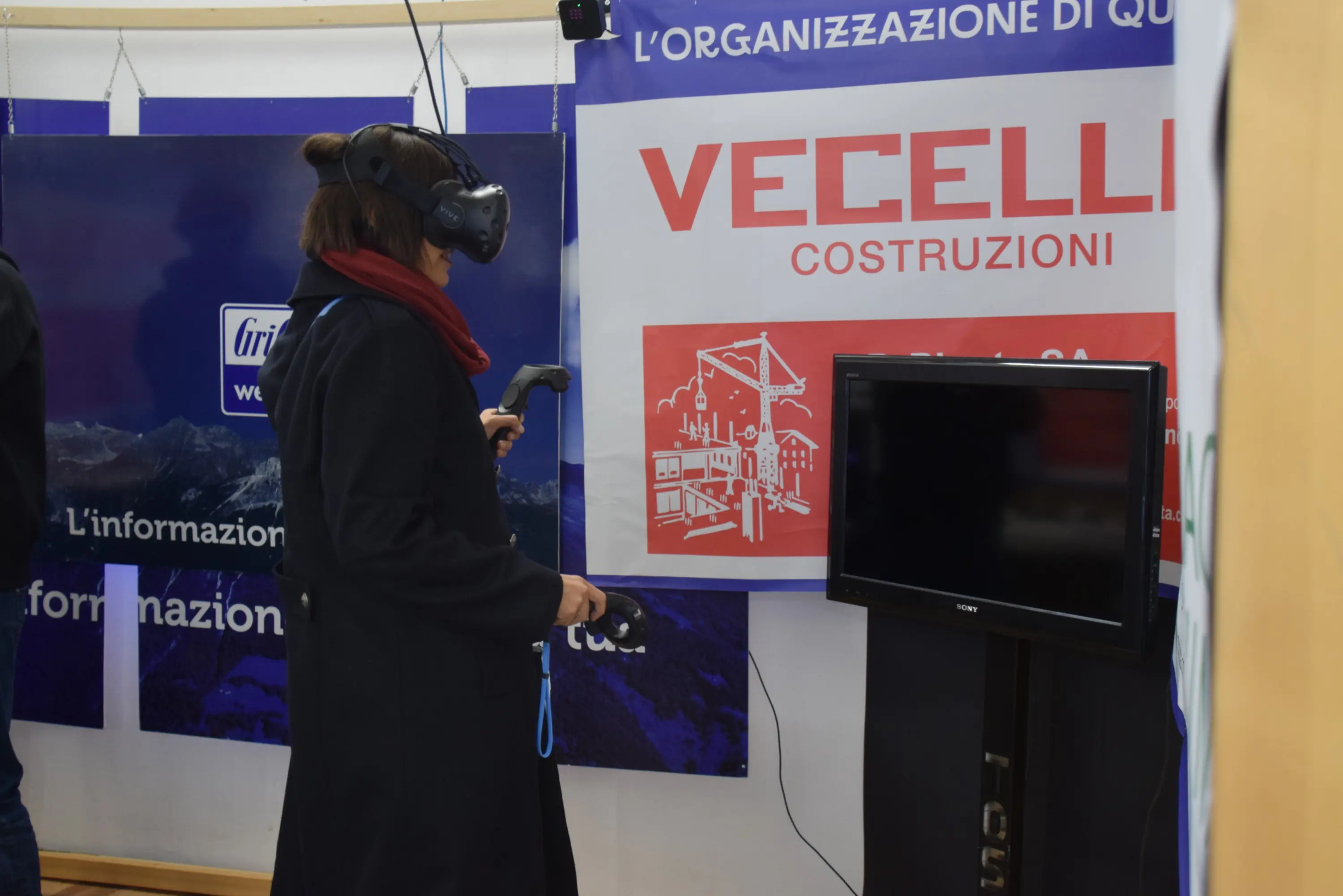Una persona indossa un visore per realtà virtuale mentre interagisce con un'esperienza virtuale, alla presenza di un cartellone pubblicitario di "Vecelli Costruzioni" e di un televisore.