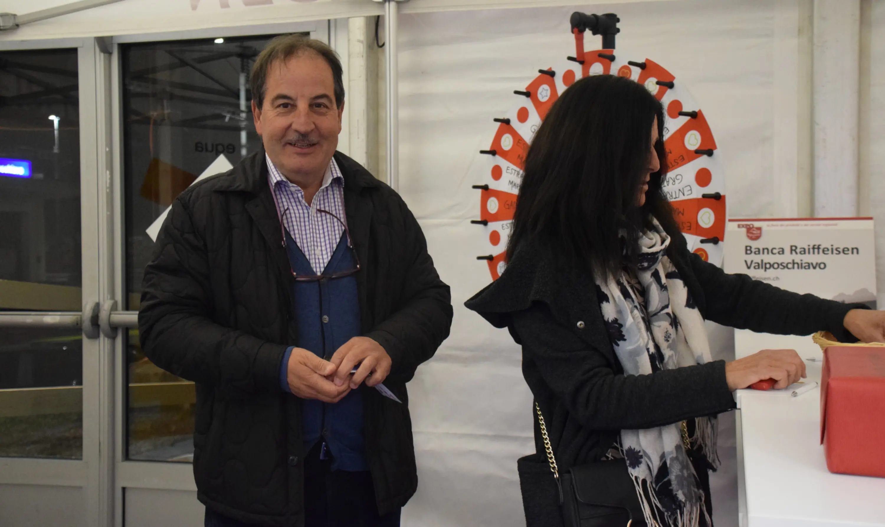 Due persone in un evento, con un uomo sorridente a sinistra e una donna con un foulard che guarda verso destra. Sullo sfondo si vede una ruota della fortuna decorata.