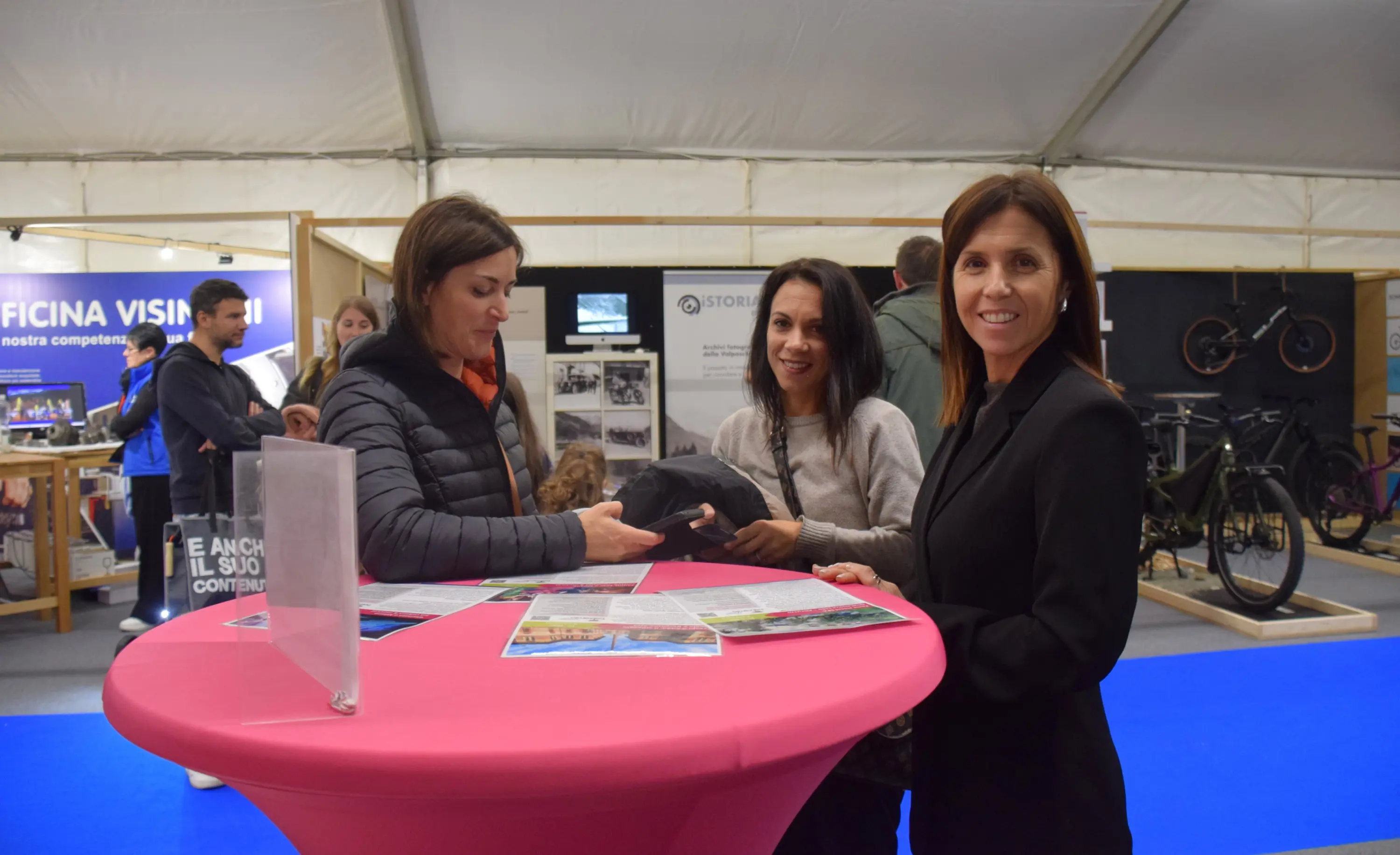 Tre donne si trovano attorno a un tavolo rosa in una fiera, mentre una di loro tiene un berretto. Sullo sfondo si vedono persone e stand espositivi.